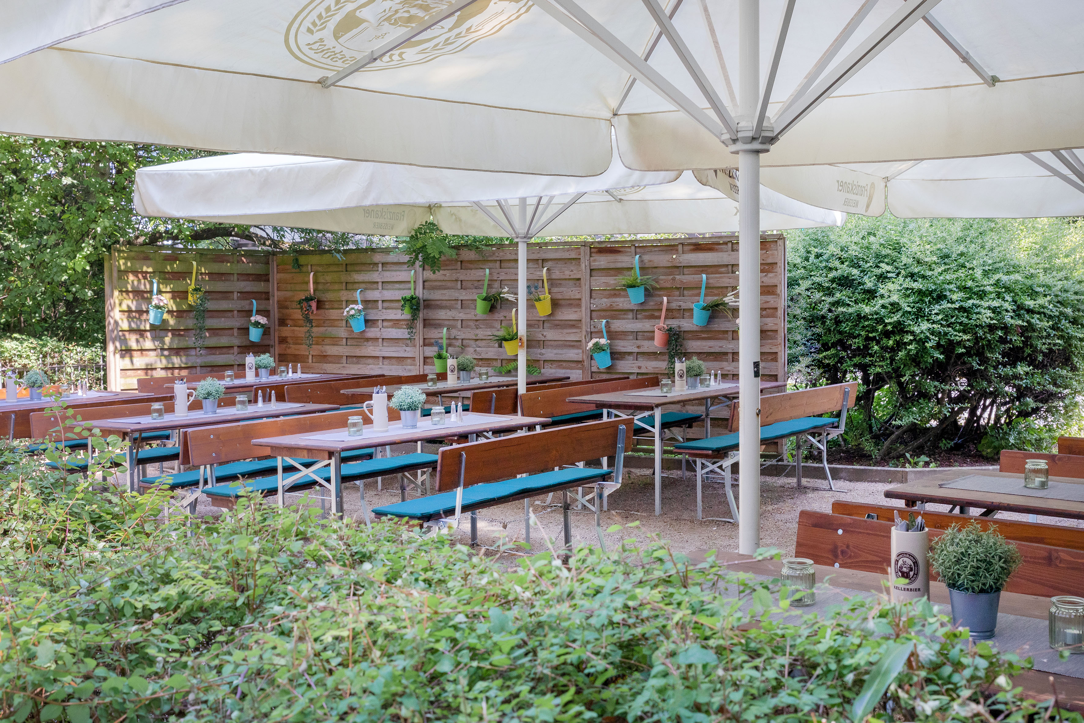 Shaded beer garden patio with wooden tables, blue benches, and green plants.