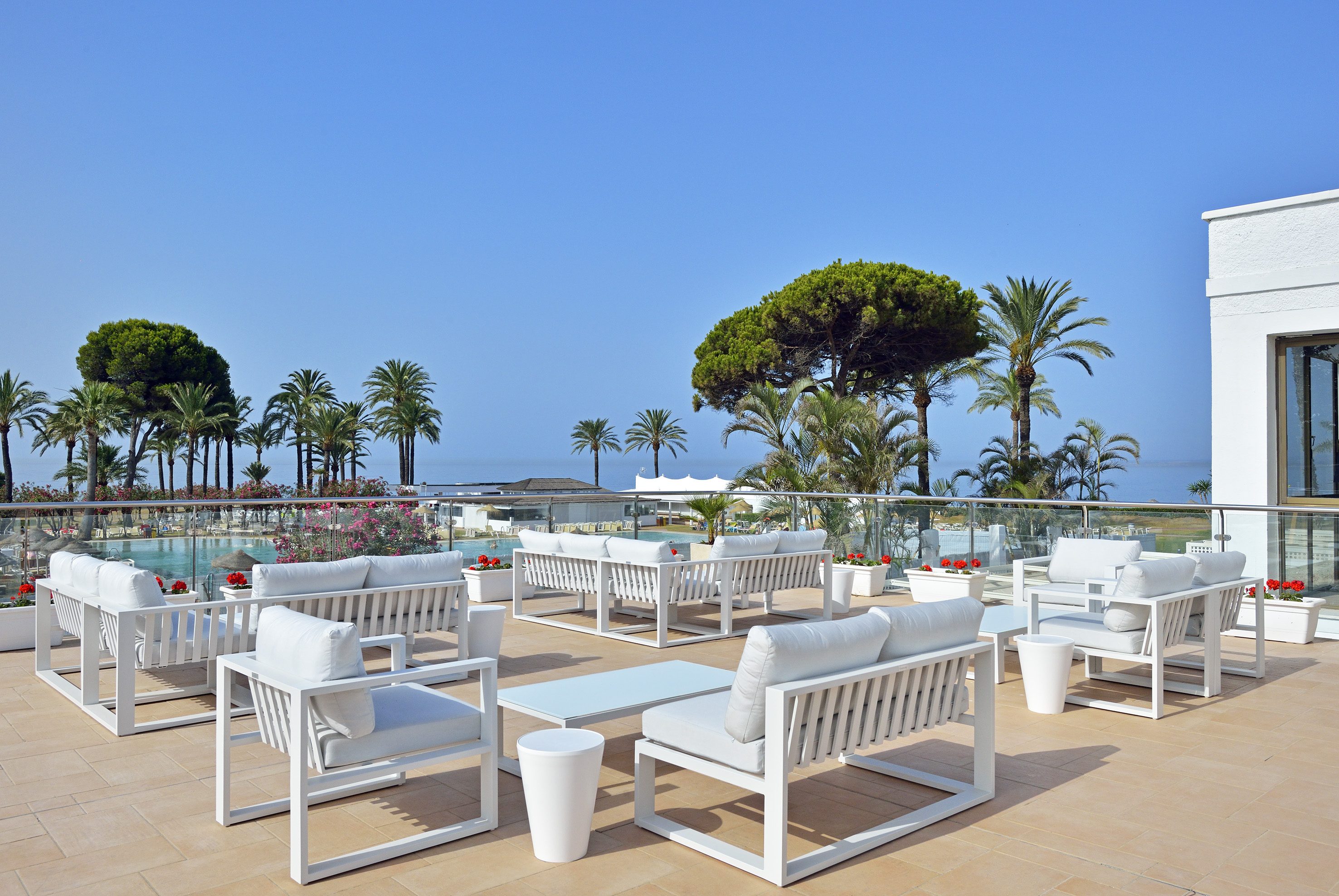 Hotel terrace lounge with white furniture, palm trees, pool, and sea view.