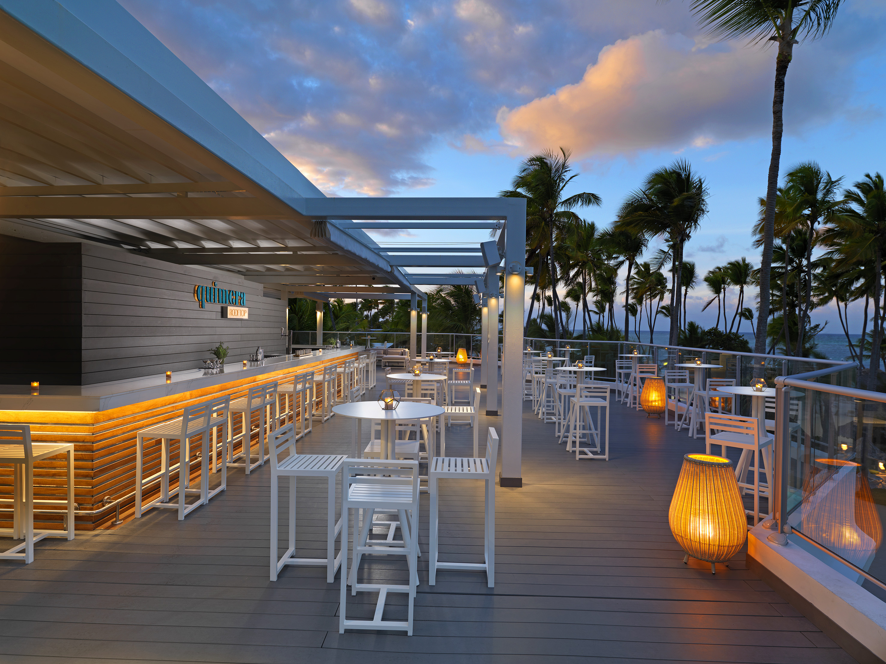 Rooftop bar at sunset, white seating, glowing counter, and palm trees.