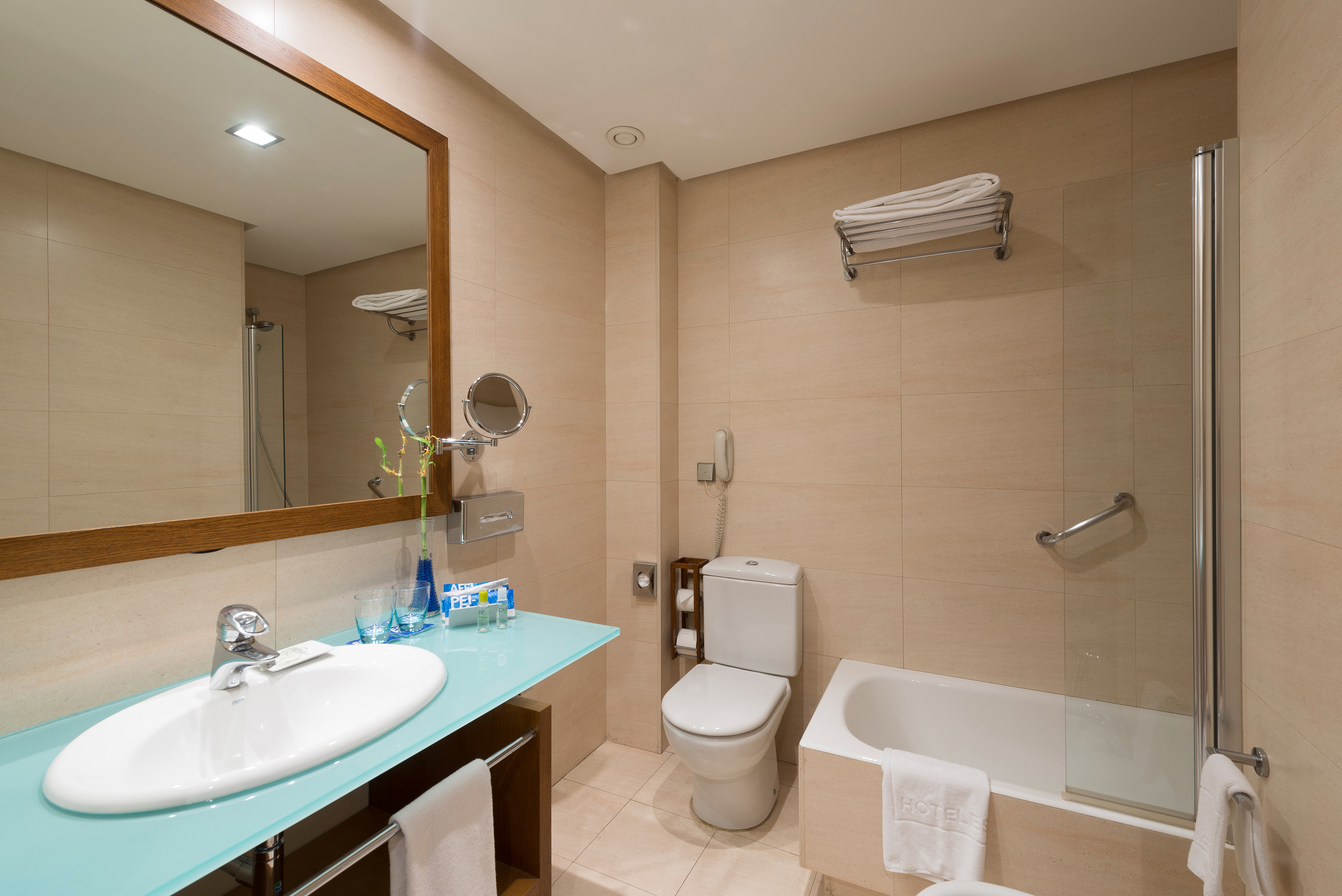Modern hotel bathroom with beige tiles, white sink, mirror, toilet, and bathtub.