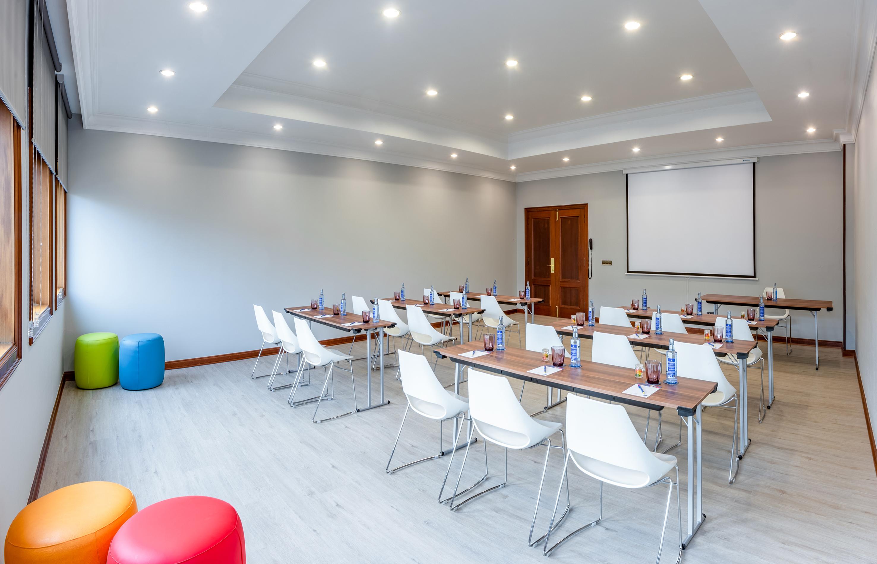 Modern meeting room with tables, white chairs, projector screen, and colorful poufs.