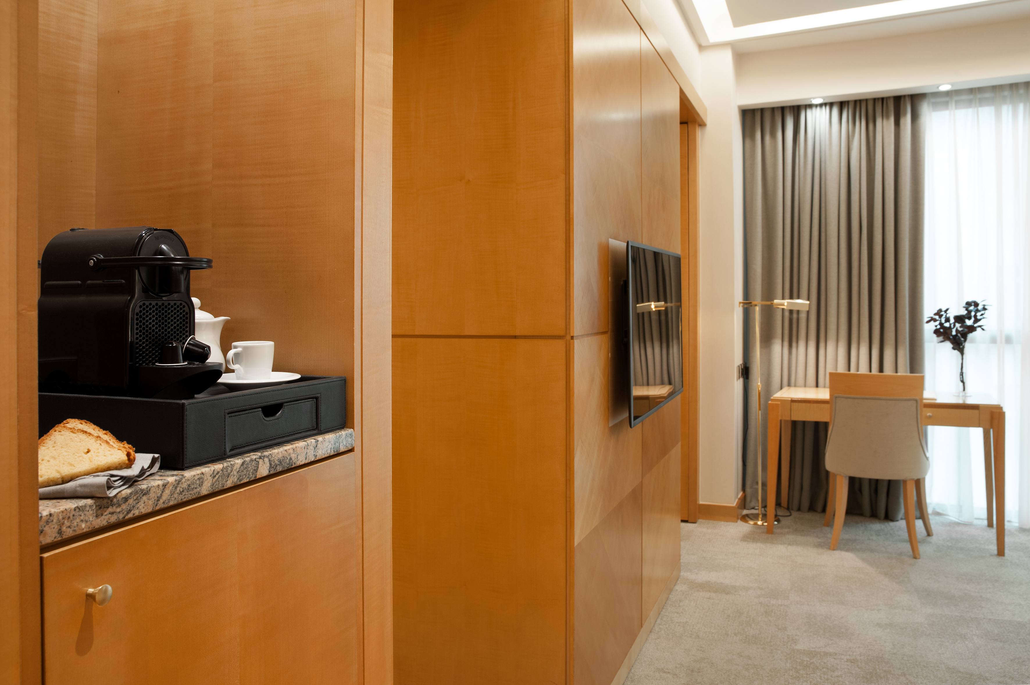 Modern hotel room with Nespresso coffee machine, wooden cabinets, desk, and TV.