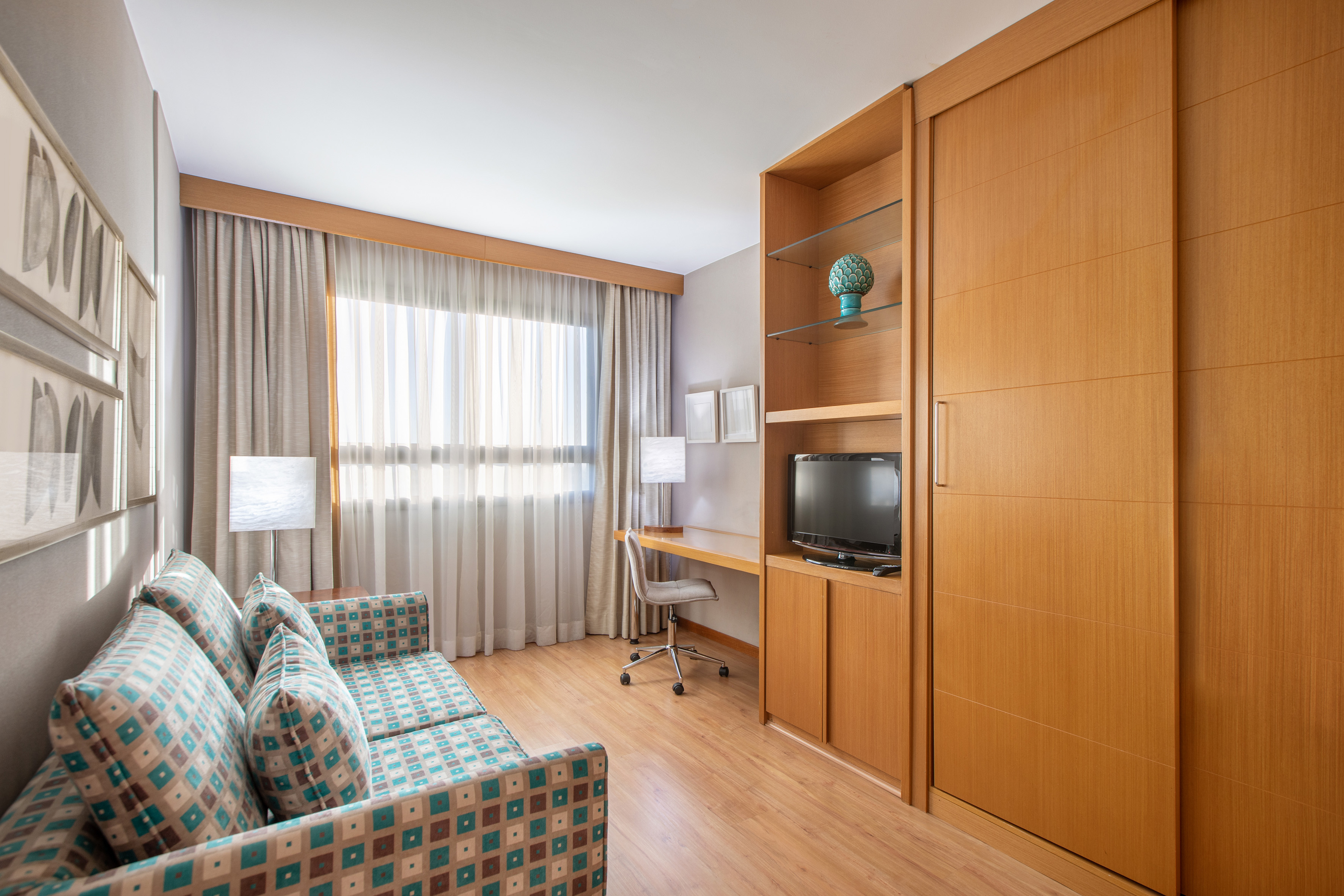 Modern hotel suite with patterned sofa, desk, TV unit, and wooden wardrobe.