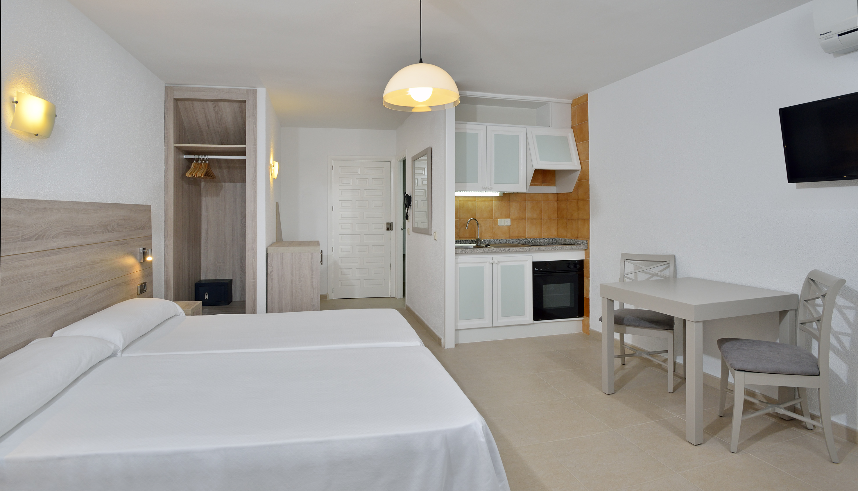 Hotel studio with twin beds, integrated kitchenette, dining table, and TV.