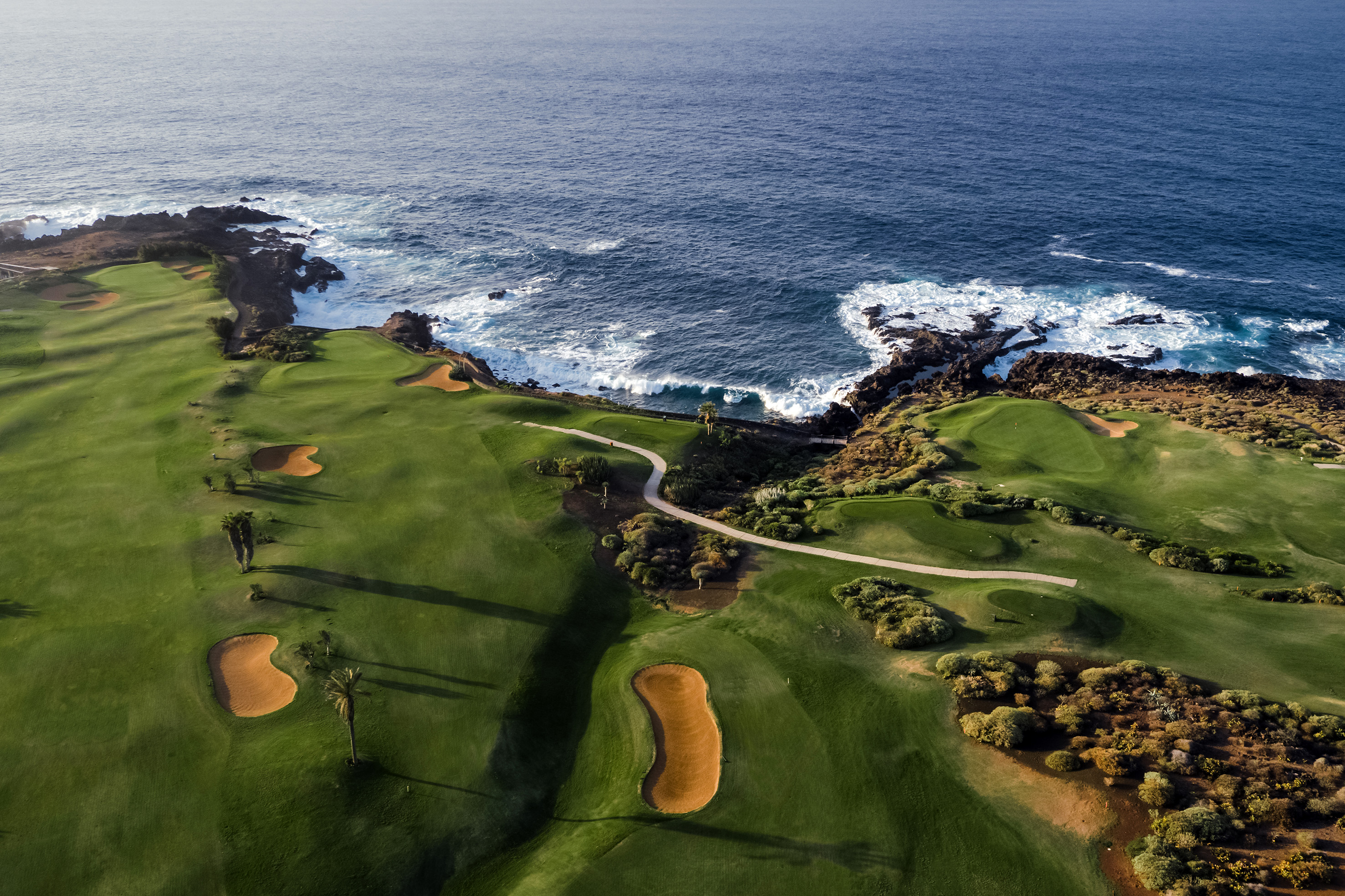 Aerial view of coastal golf course with green fairways, bunkers, and ocean waves.