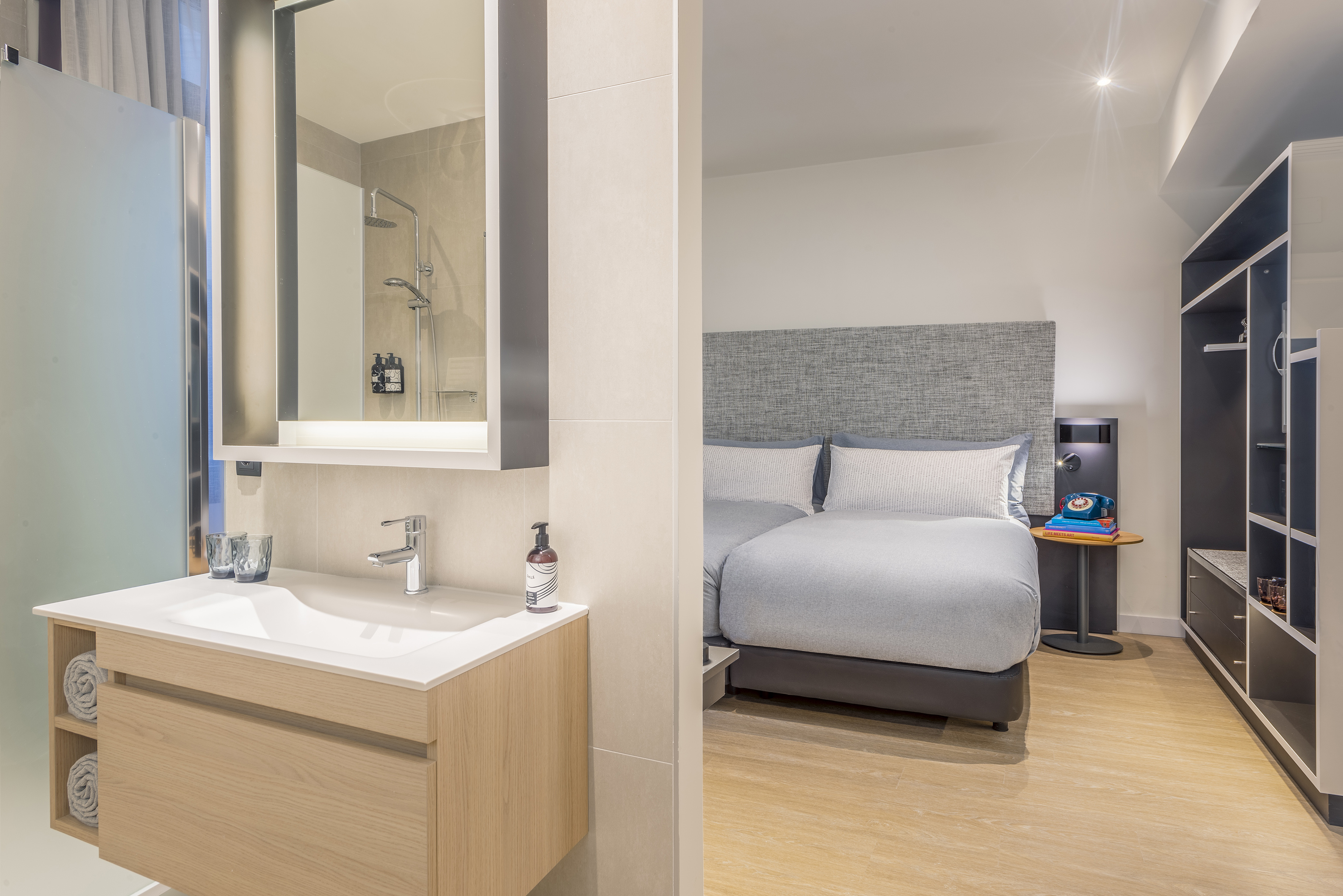 Modern hotel room interior with connected bathroom vanity and grey bed.