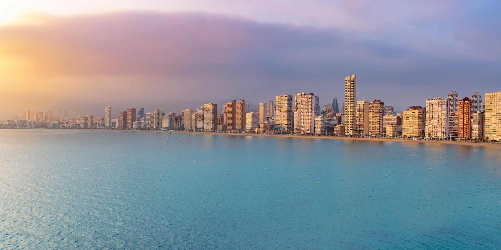 Coastal city skyline with modern buildings, vast ocean, and warm sunset glow.