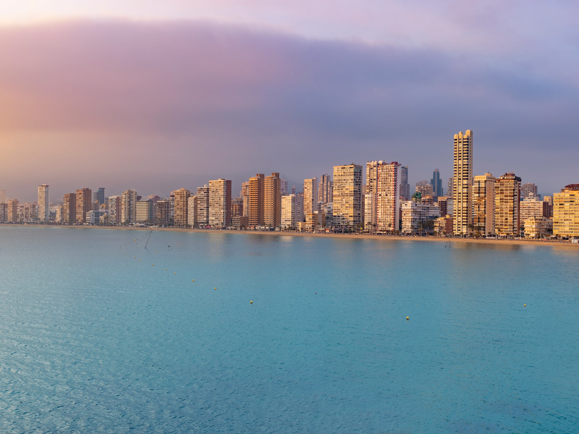 Dense urban skyline along a wide beach and calm sea, hazy morning light.