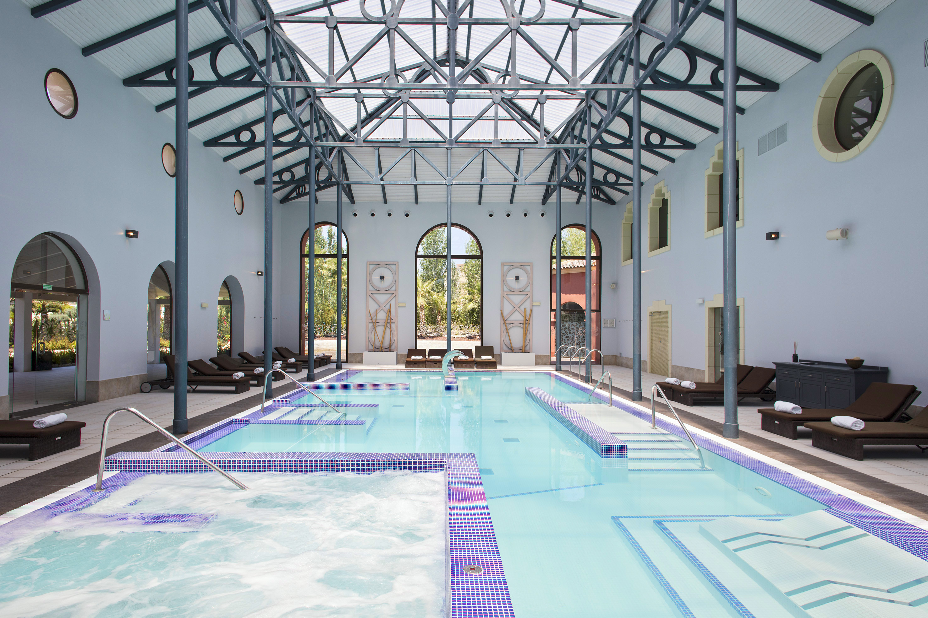 Luxury indoor pool with architectural glass roof, jacuzzi, and lounge seating.