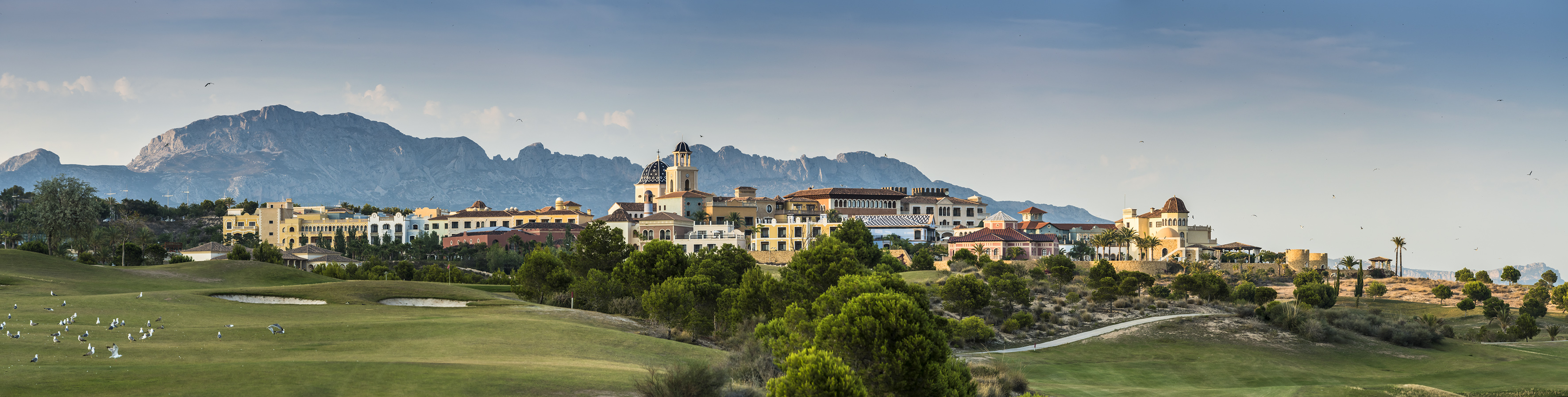 Mediterranean village resort with golf course and mountains under blue sky.
