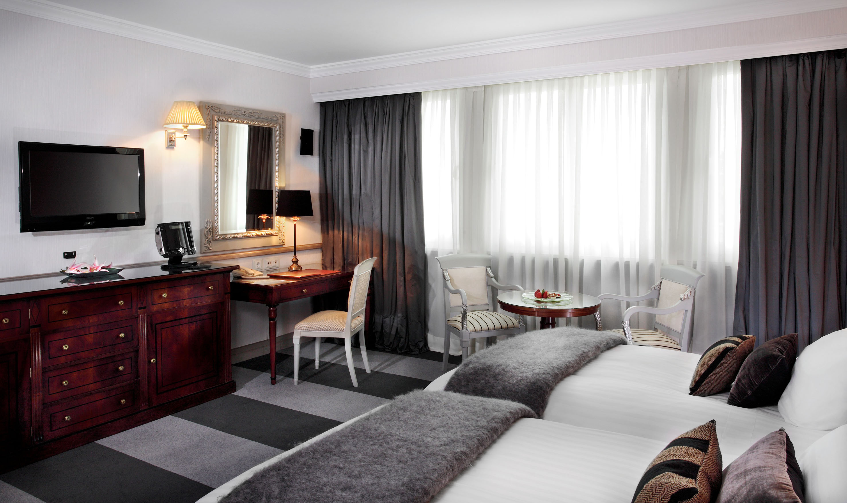 Luxury hotel bedroom with twin beds, workspace, TV, and window.