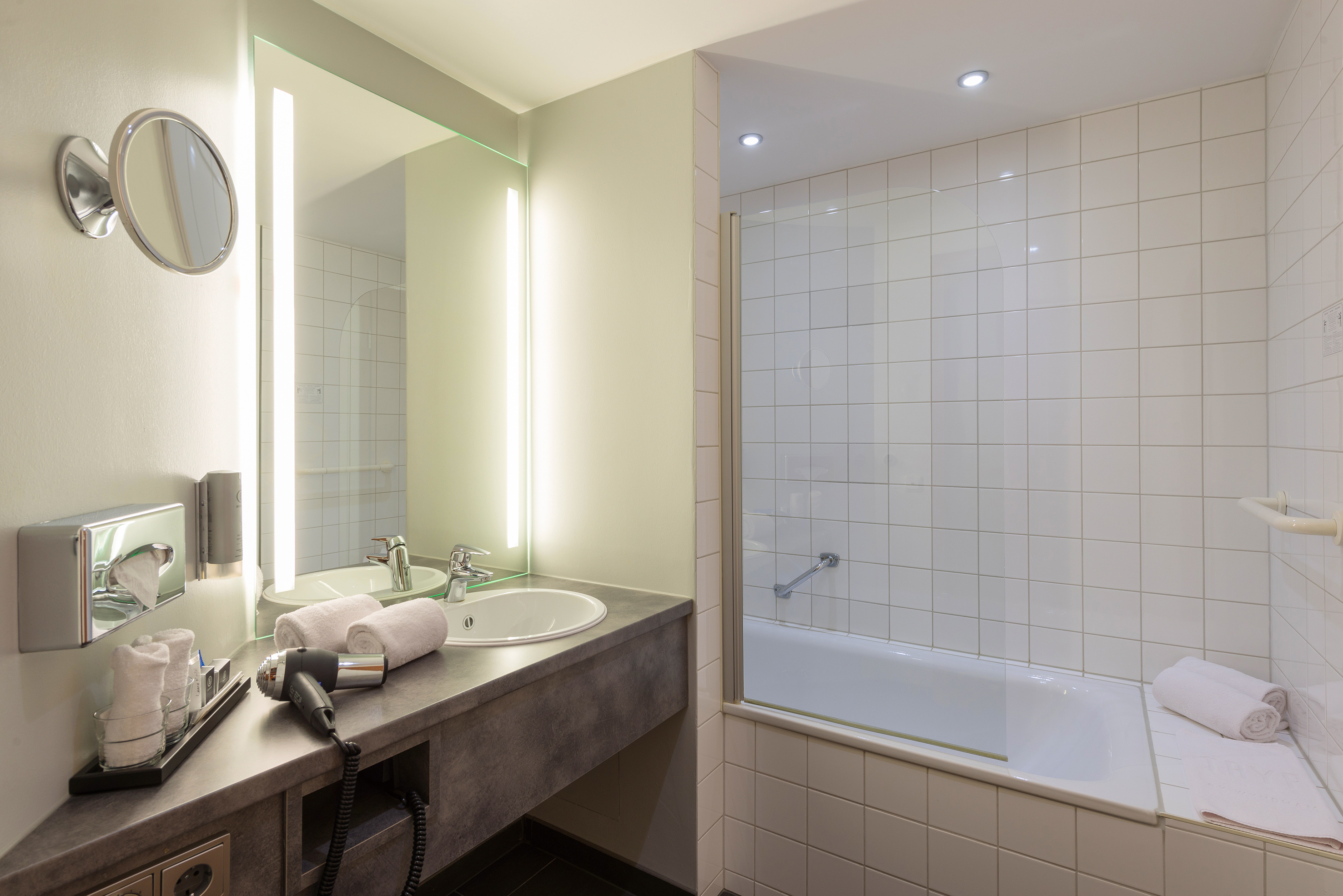 Modern bathroom with sink, lighted mirror, hairdryer, and white tiled bathtub.