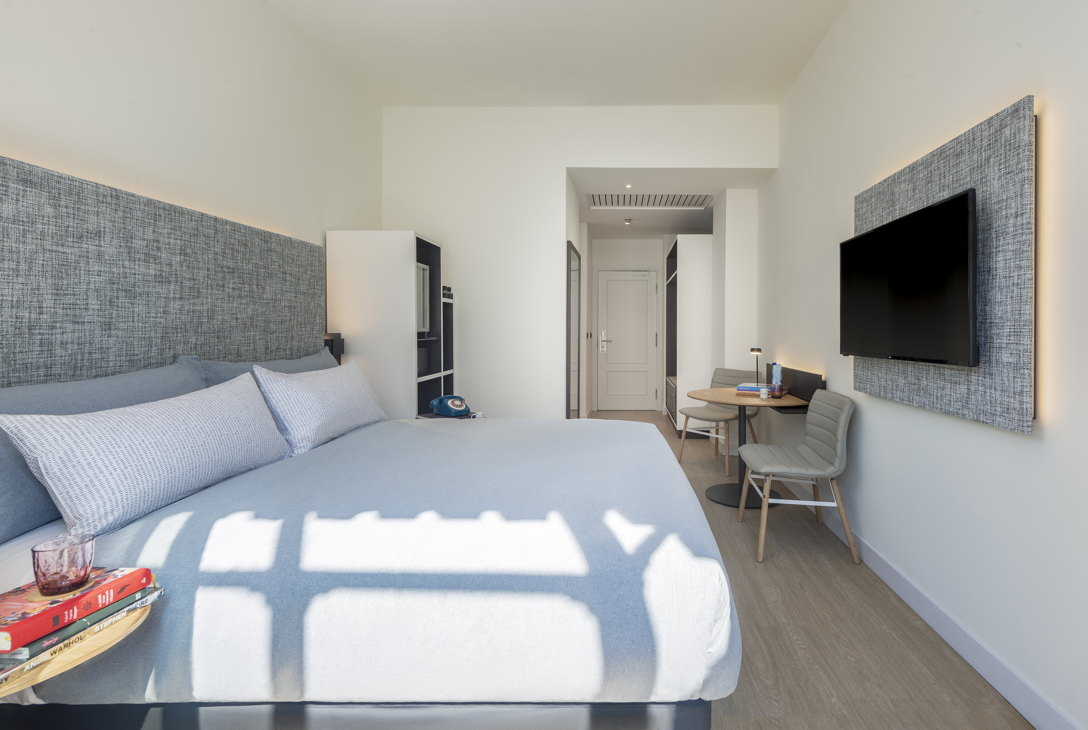 Modern hotel room with light blue bed, LED headboard, wall TV, and small table.