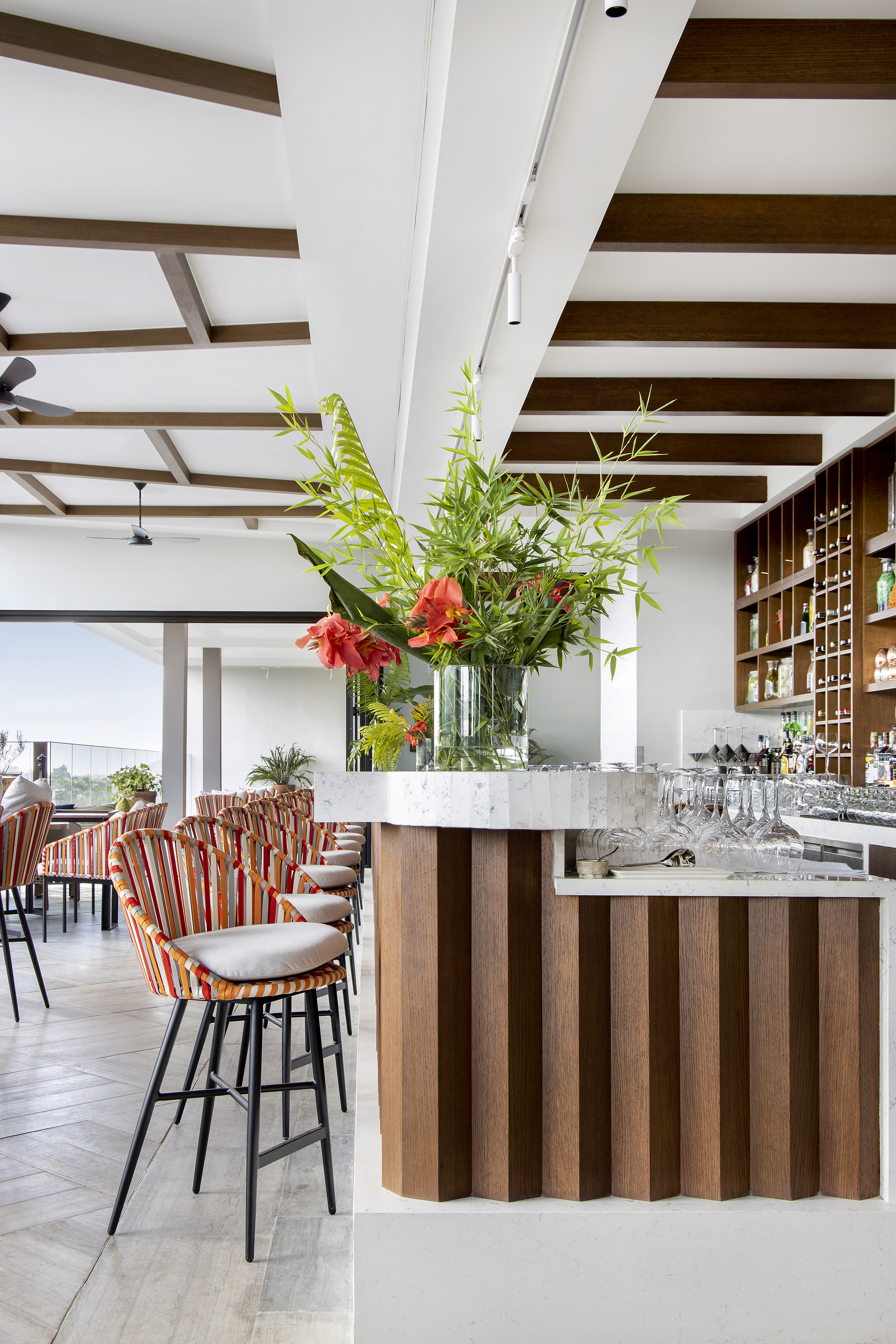 Modern bar with wooden counter, marble top, striped stools, and tropical flowers.