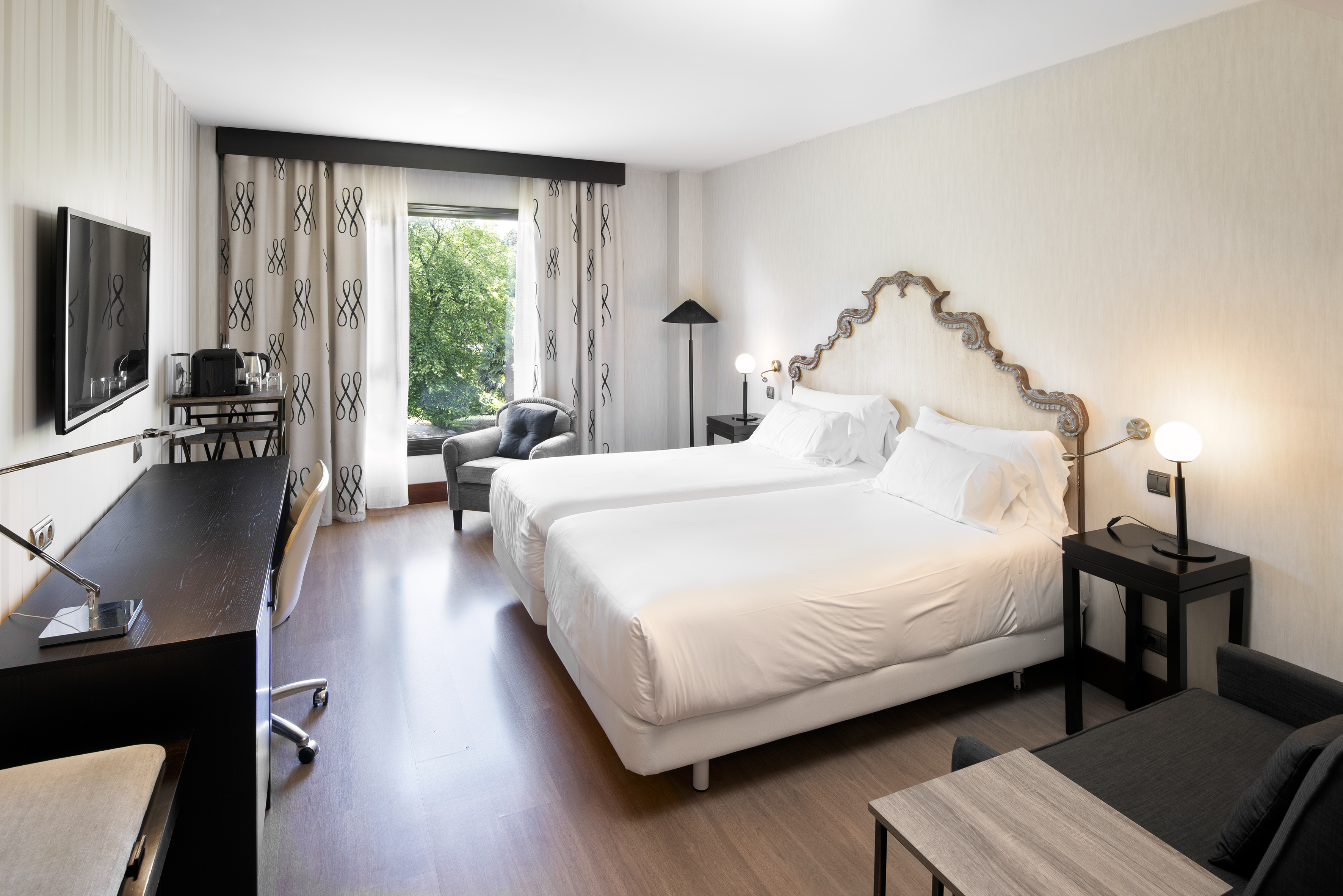Modern hotel room with twin beds, desk, and large window overlooking trees.