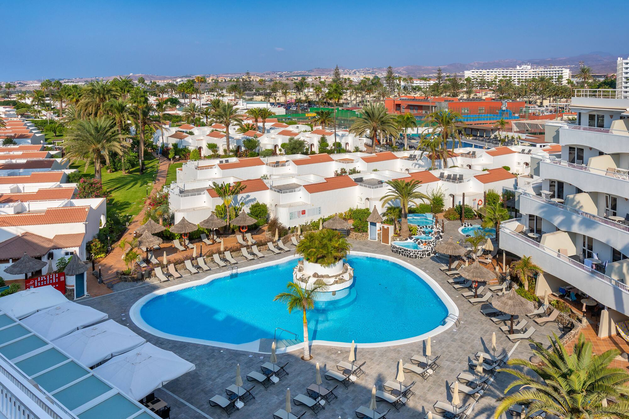 Aerial view of a tropical resort with a large swimming pool, palm trees, and white bungalows.