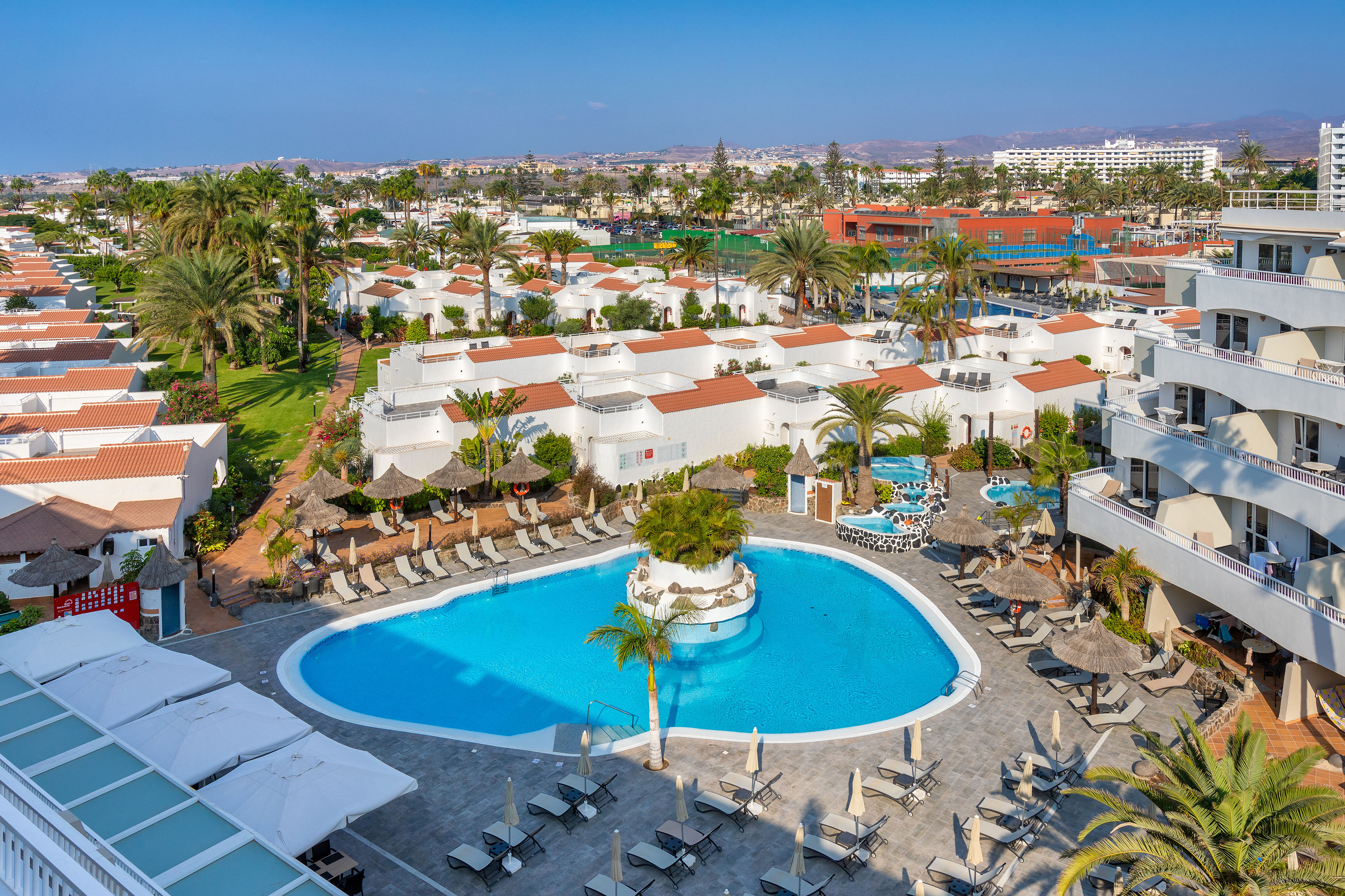 Aerial view of a tropical resort with a large swimming pool, palm trees, and white bungalows.