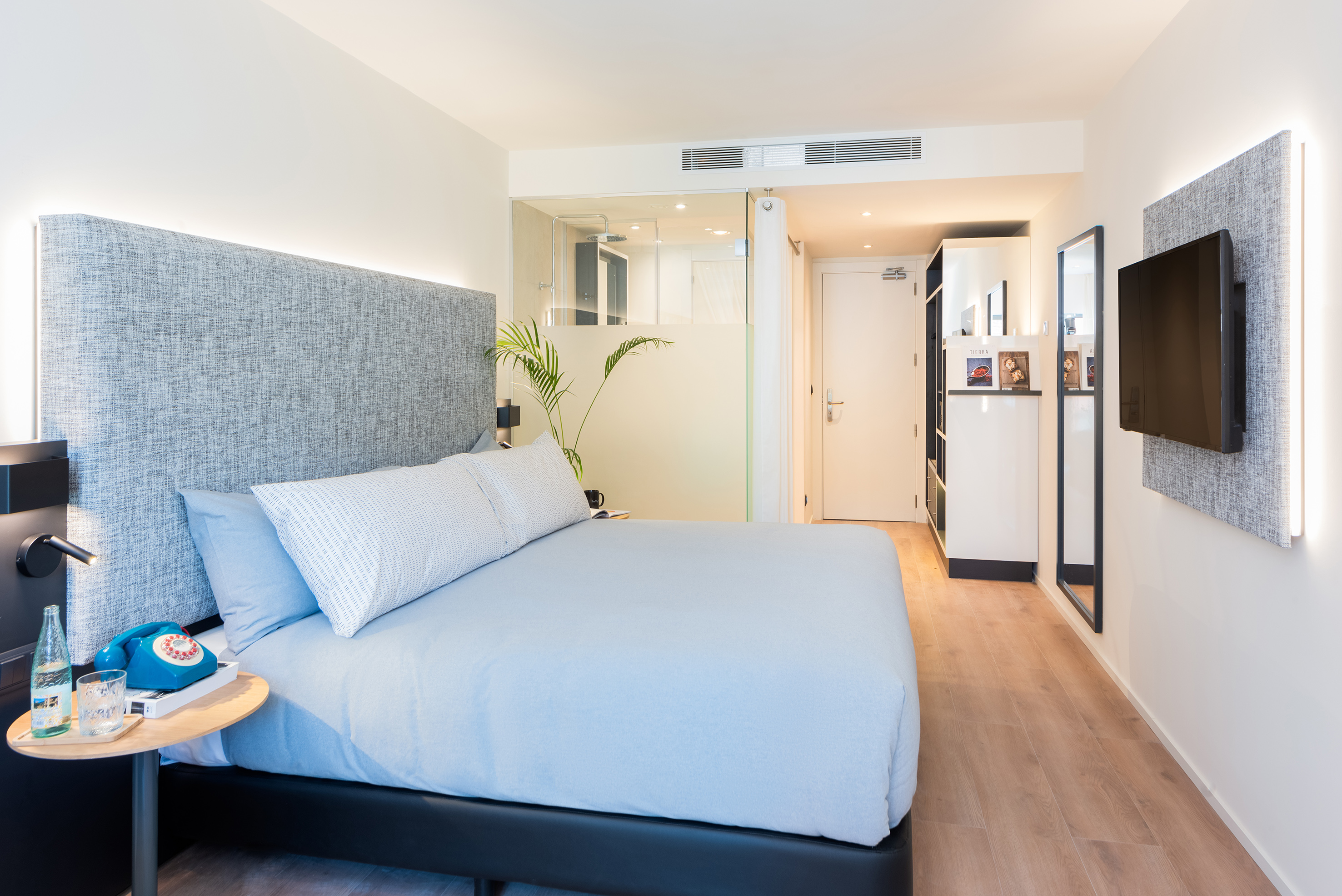Modern hotel bedroom with light blue bed, backlit headboard, and glass shower.