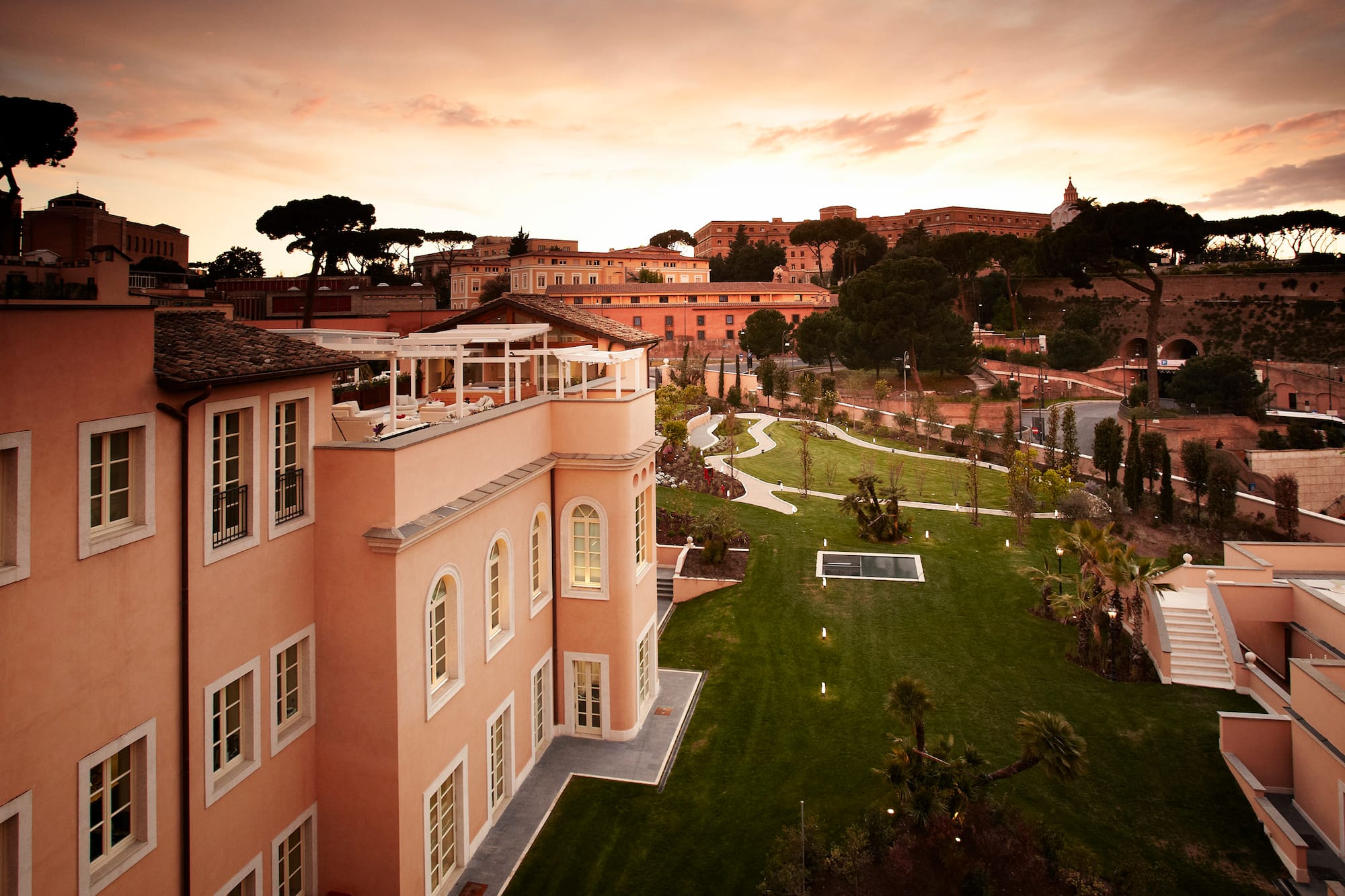 Roman hotel with rooftop terrace, lush gardens, and cityscape at sunset.