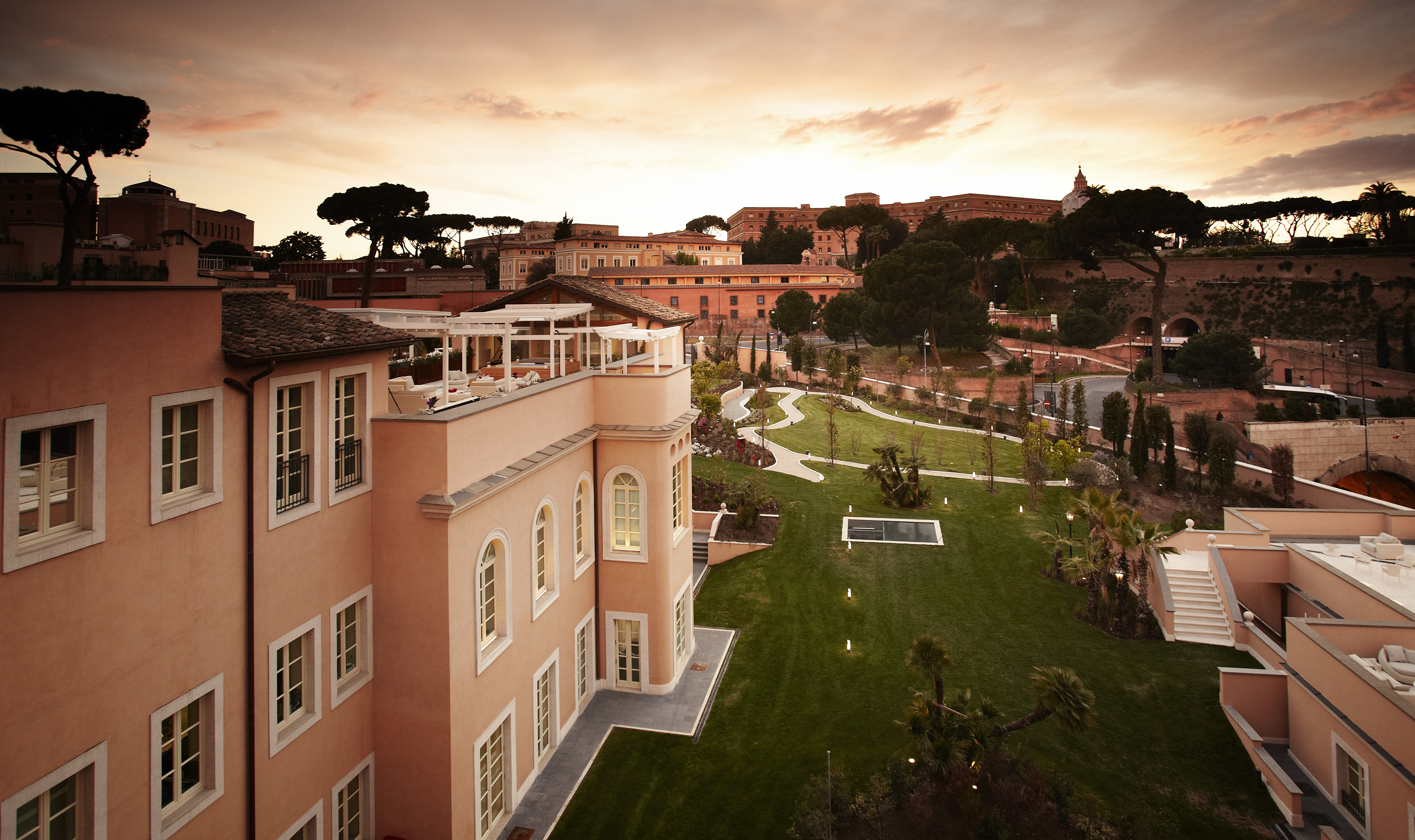 Roman hotel with rooftop terrace, lush gardens, and cityscape at sunset.