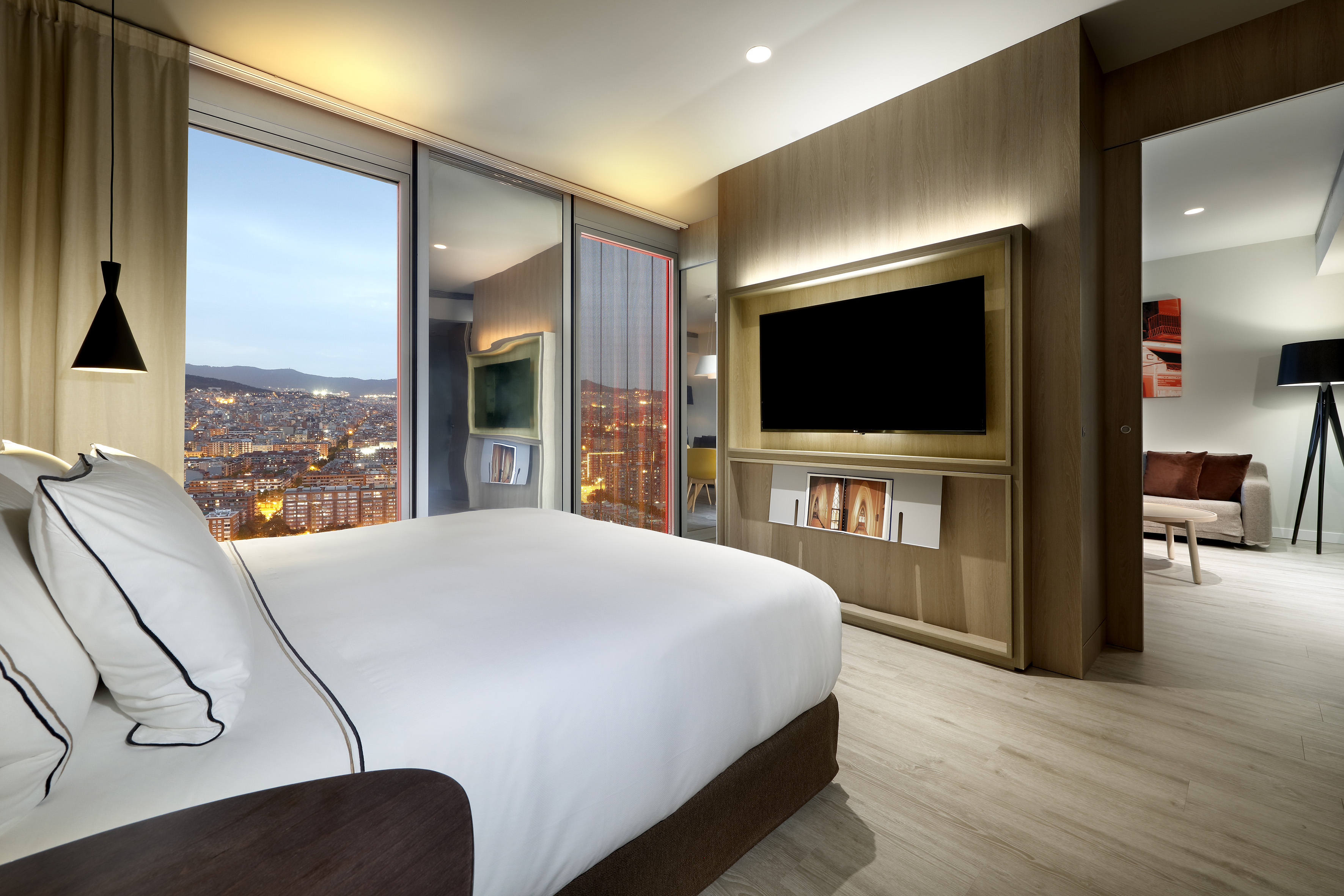 Modern hotel bedroom with city skyline view at dusk. King bed, wall-mounted TV.