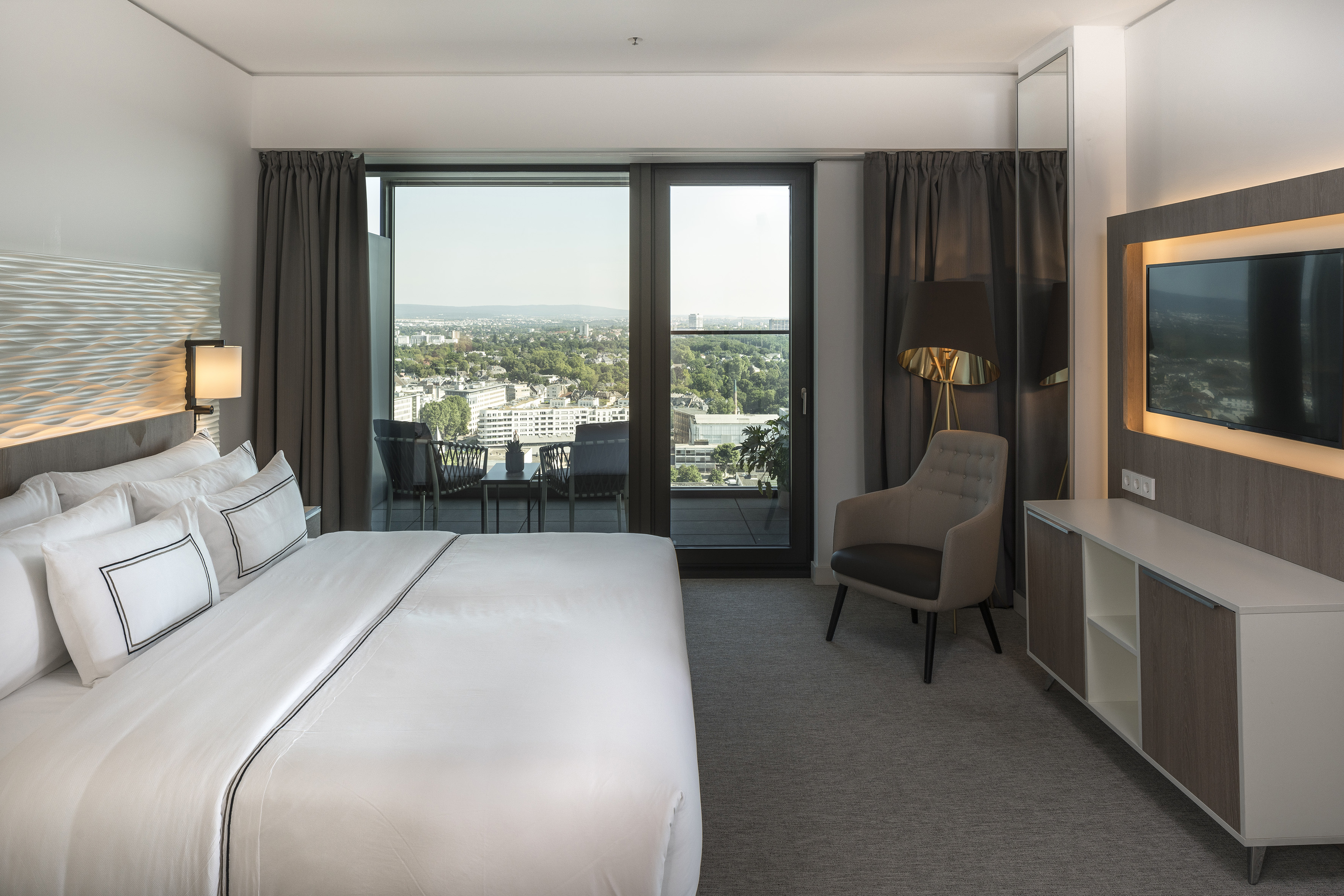 Modern hotel room with large bed, city view balcony, and contemporary design.