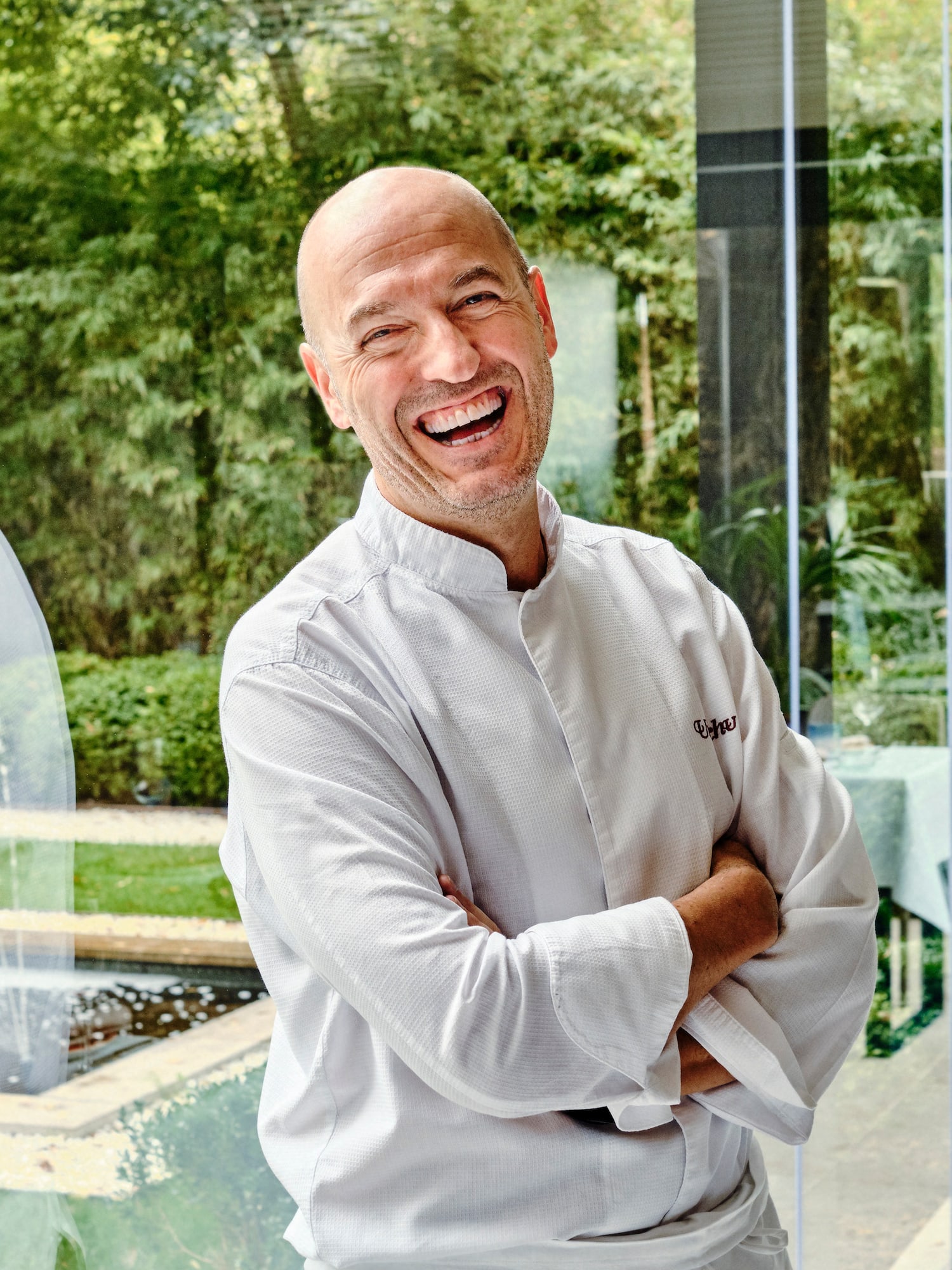 Chef Uku smiling in white uniform, arms crossed, with green outdoor background.