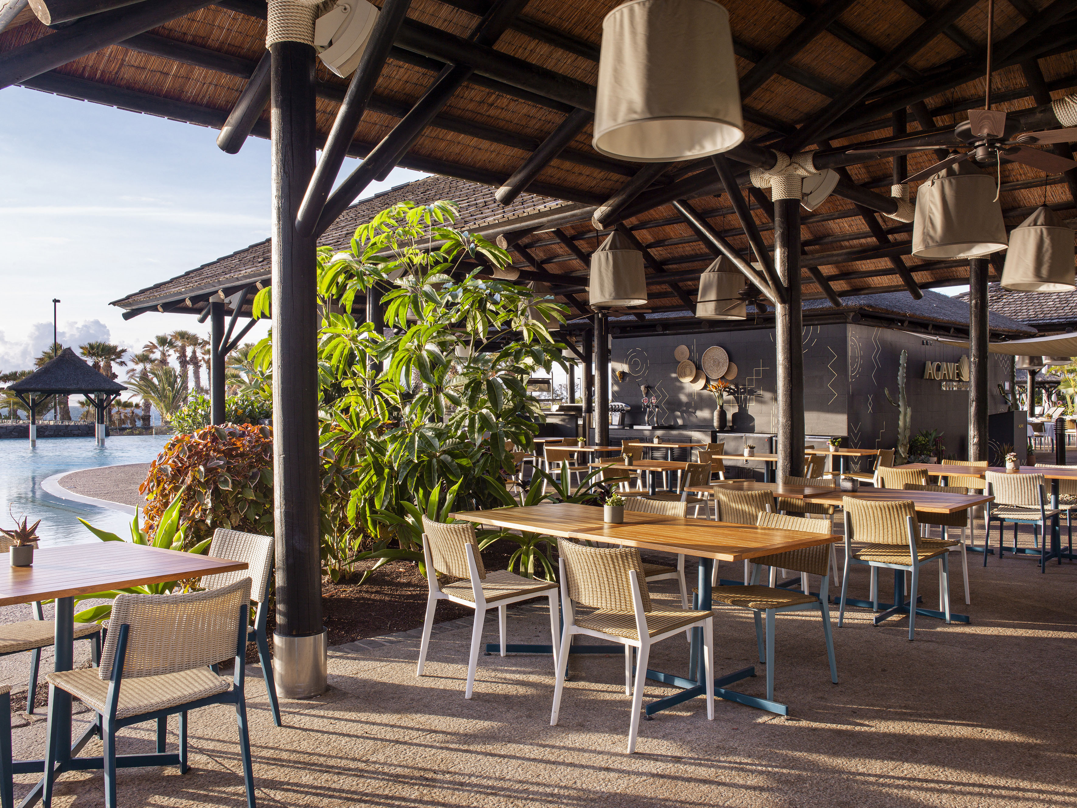 Outdoor restaurant with thatched roof, wood tables, woven chairs, and poolside view.