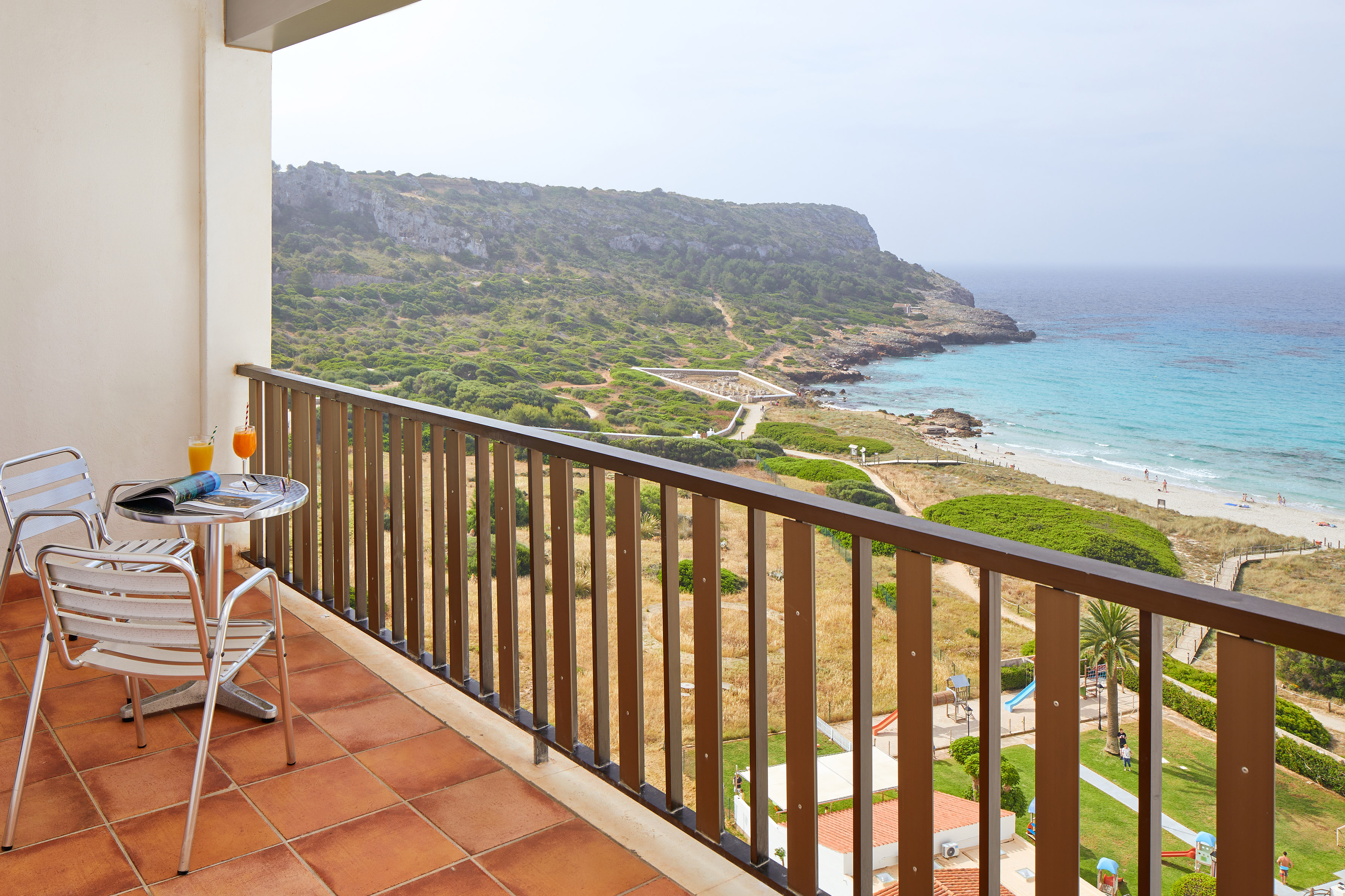 Balcony with chairs, drinks, scenic sandy beach, turquoise sea, and coastal cliffs.
