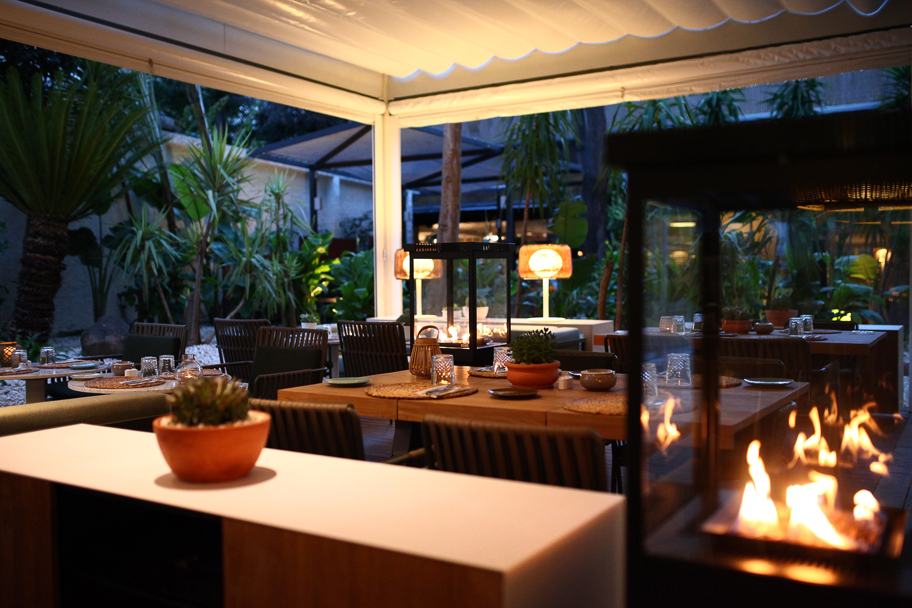 Outdoor dining patio with tables, tropical plants, and warm fire features at night.