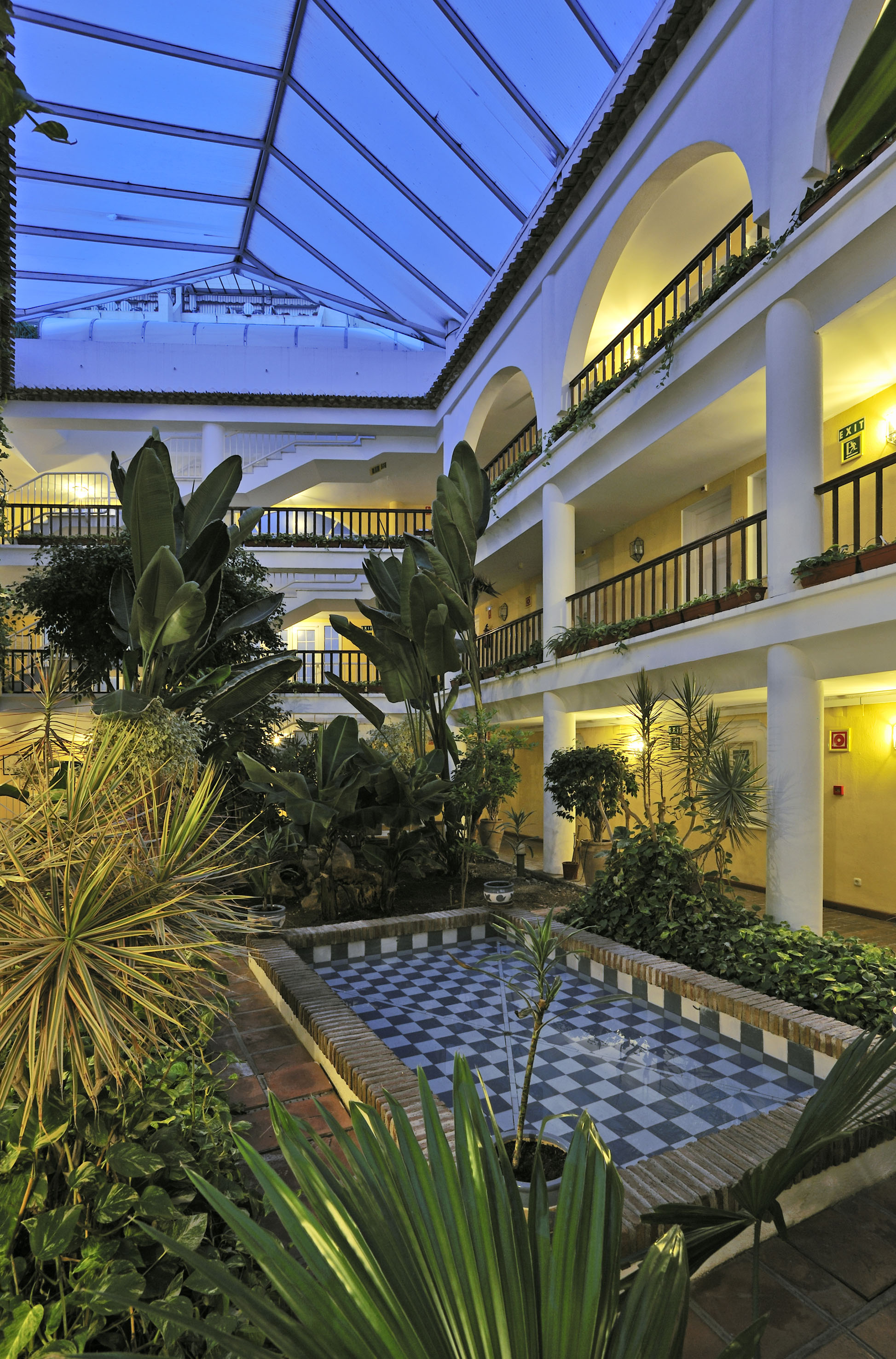 Hotel atrium with lush tropical garden, arched balconies, and checkered fountain.