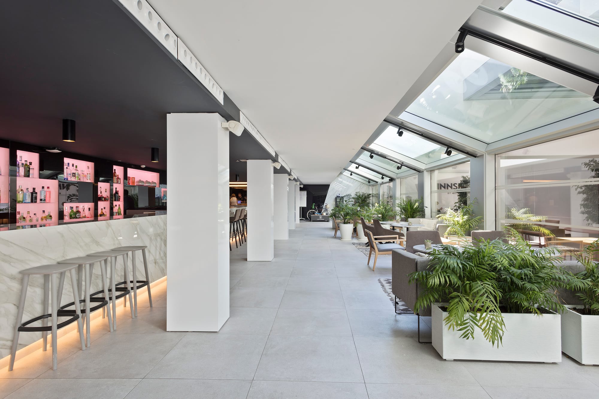 Modern hotel bar and lounge with marble counter, bar stools, glass ceiling, and plants.