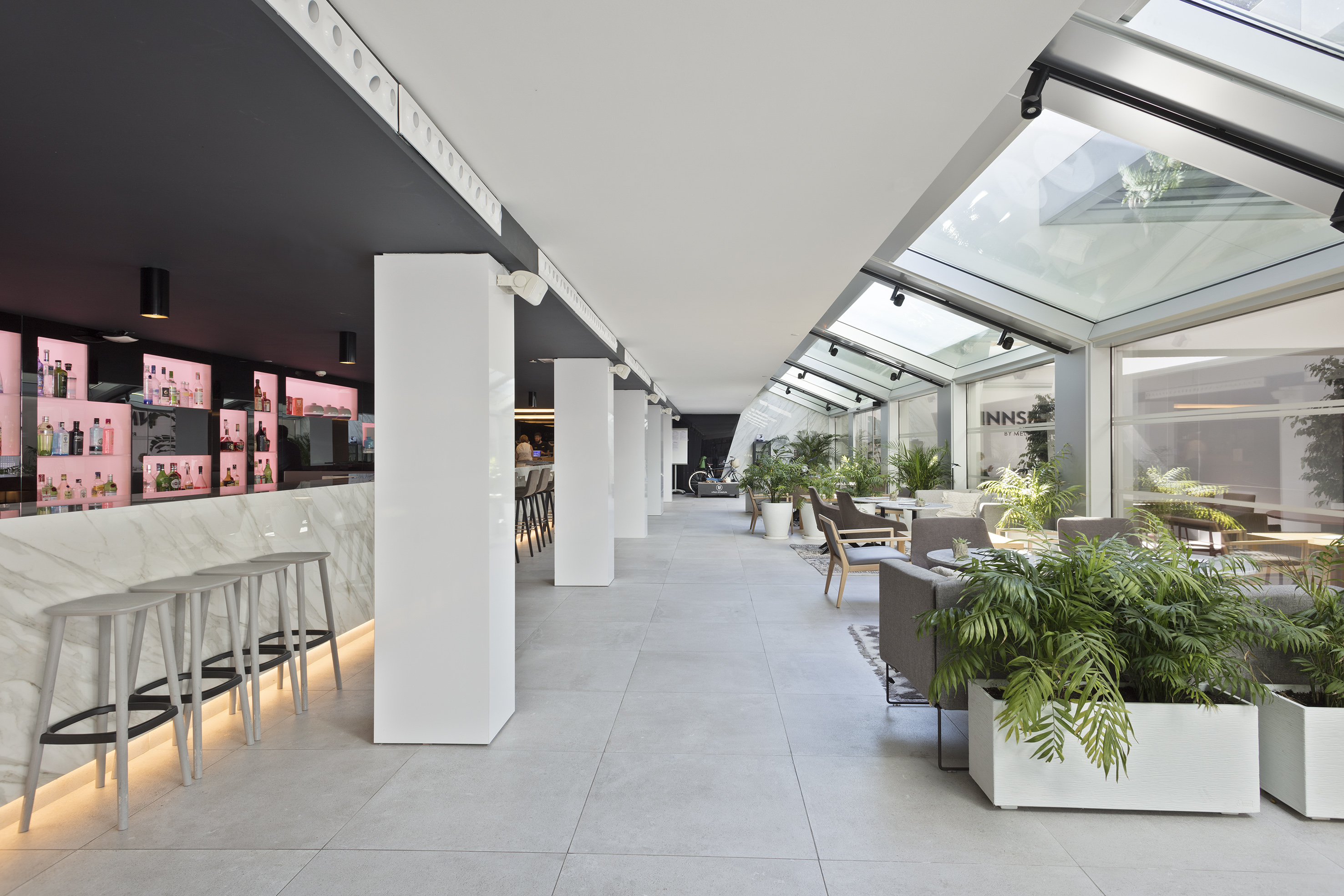 Modern hotel bar and lounge with marble counter, bar stools, glass ceiling, and plants.