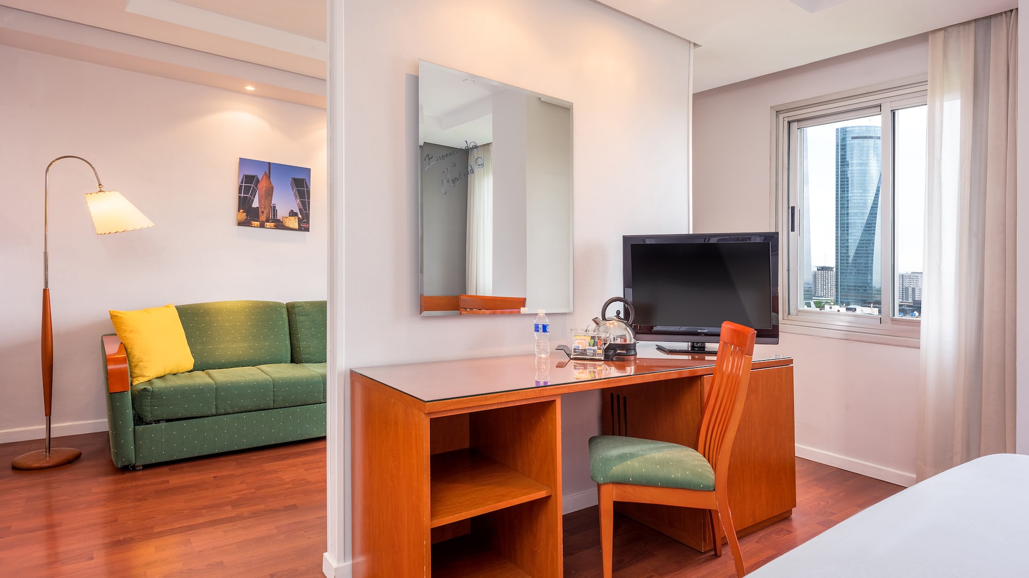 Modern hotel suite with workspace, TV, sofa, and city view window.