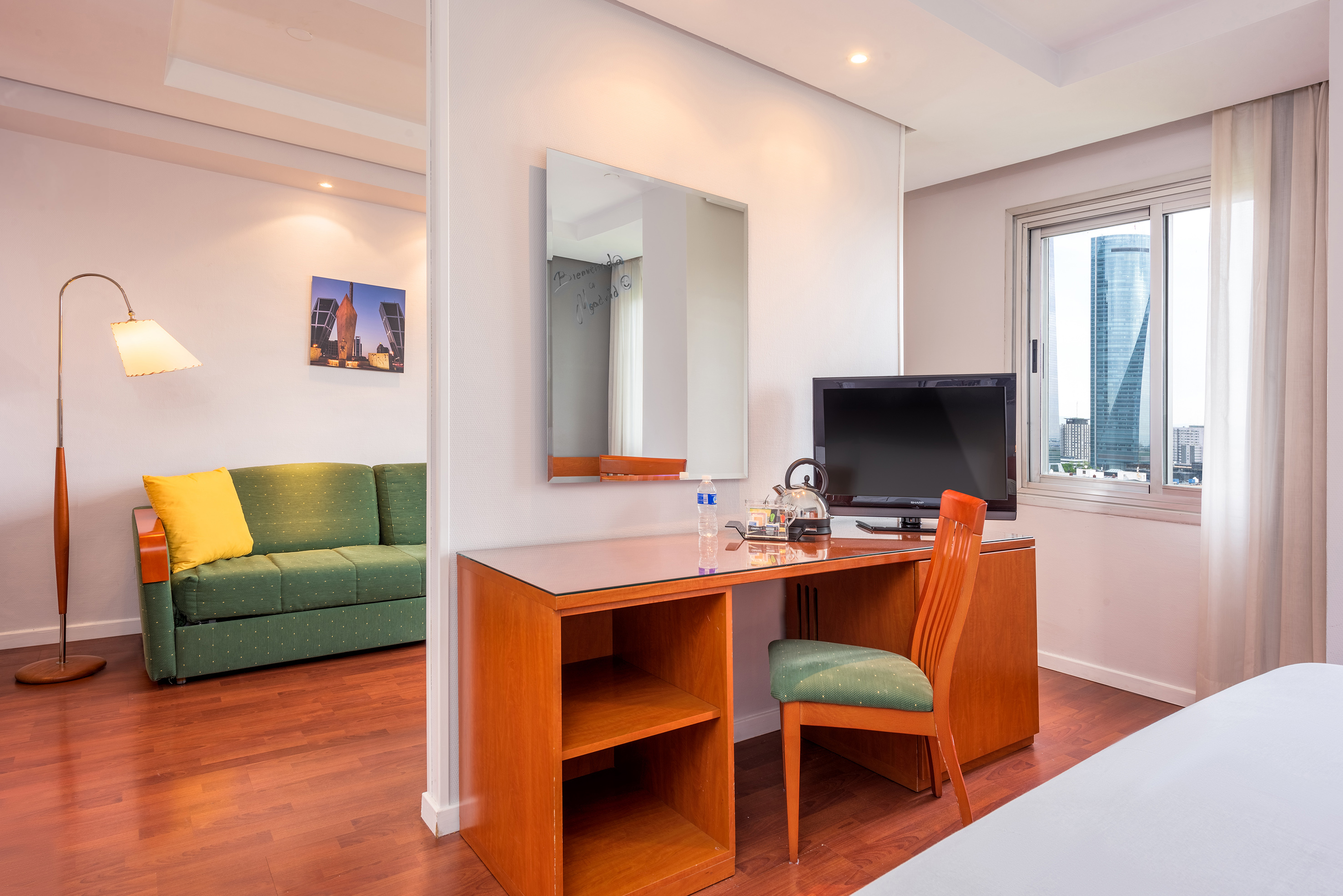 Modern hotel suite with workspace, TV, sofa, and city view window.