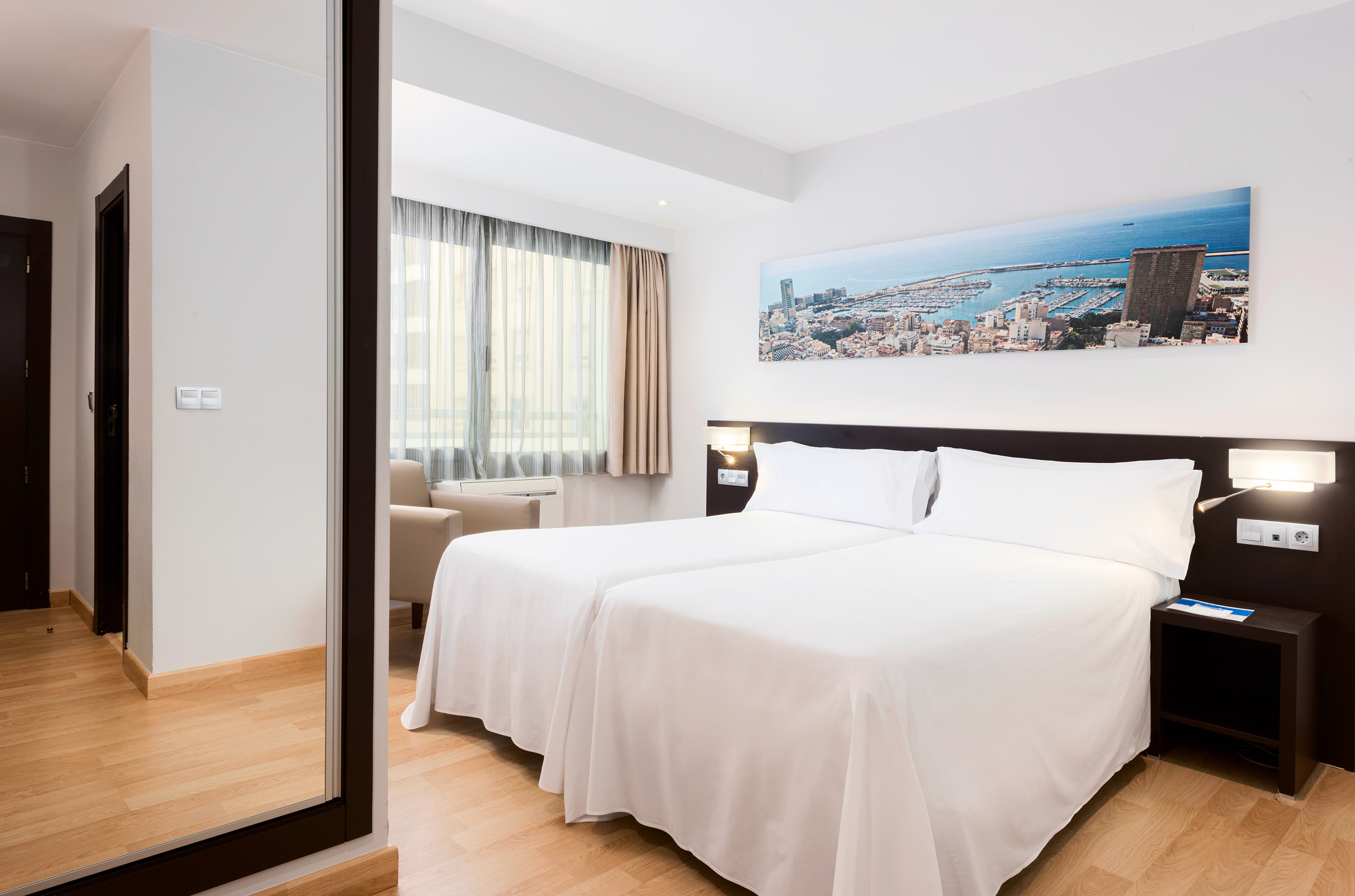 Modern hotel room with twin beds, cityscape art, window, and wood floor.