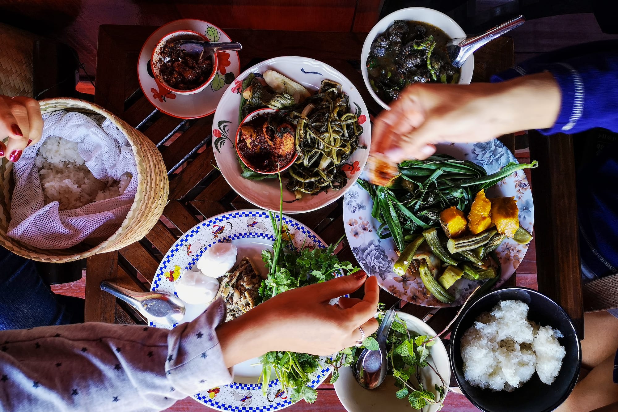 Shared Lao sticky rice meal with greens, chili paste, stew, and diners' hands.