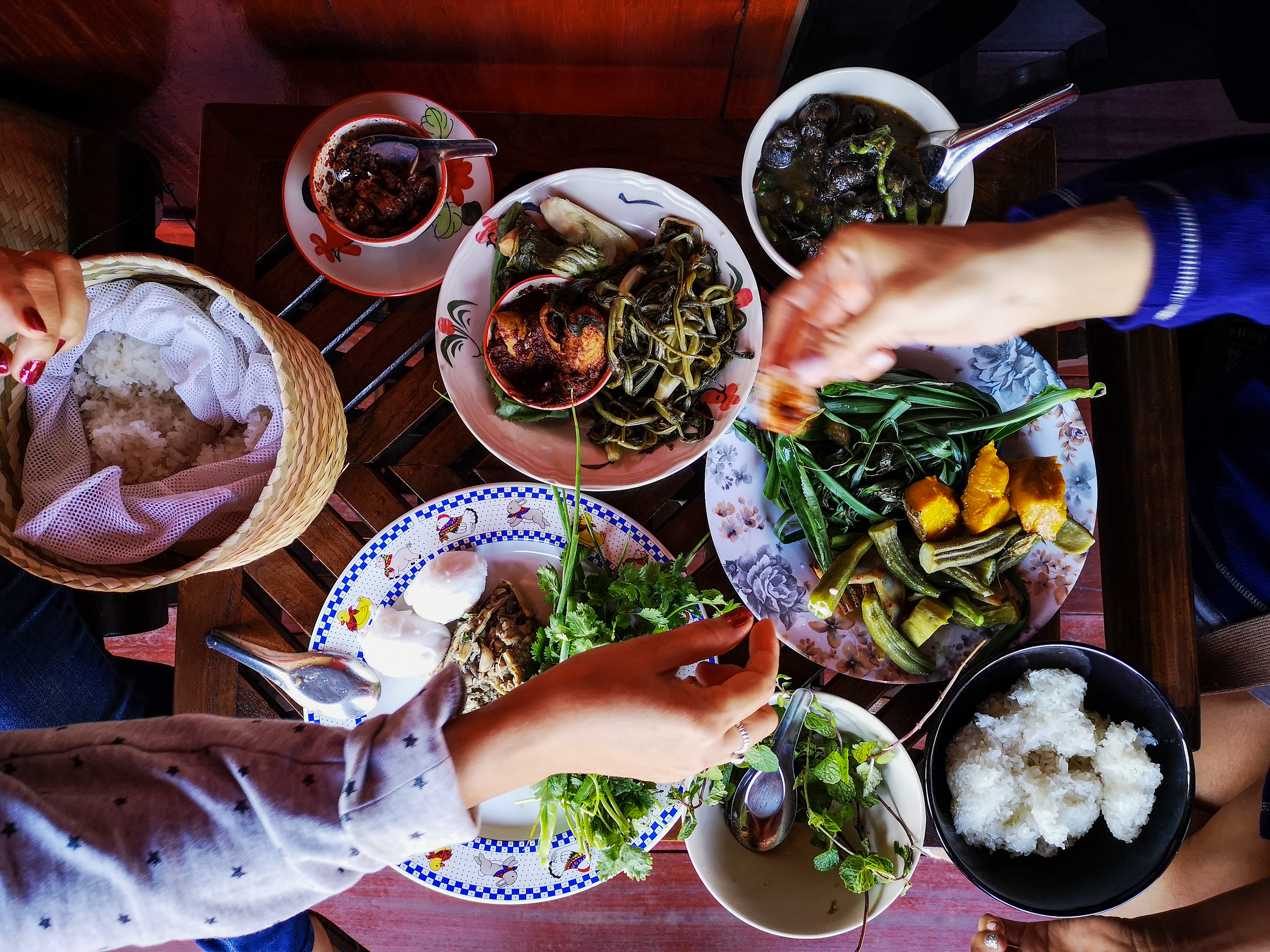Shared Lao sticky rice meal with greens, chili paste, stew, and diners' hands.