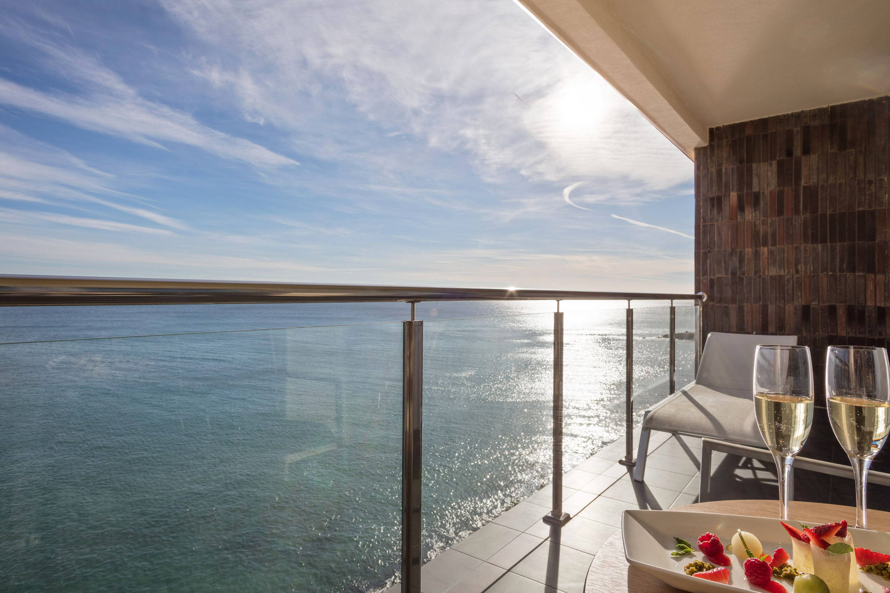 Sunny ocean view balcony with champagne, fruit dessert, and glass railing.