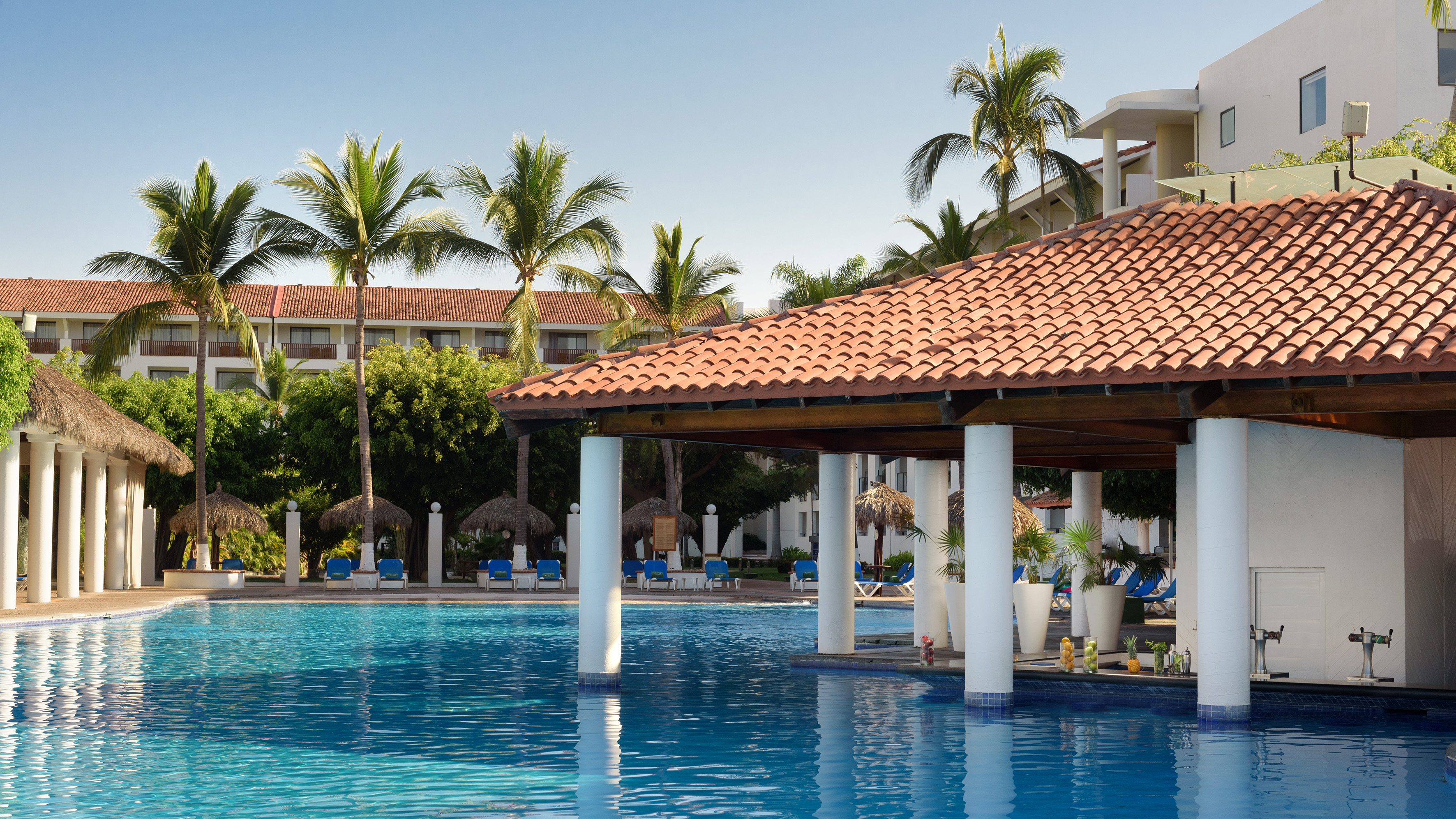 Tropical resort pool with swim-up bar, palm trees, and hotel buildings.