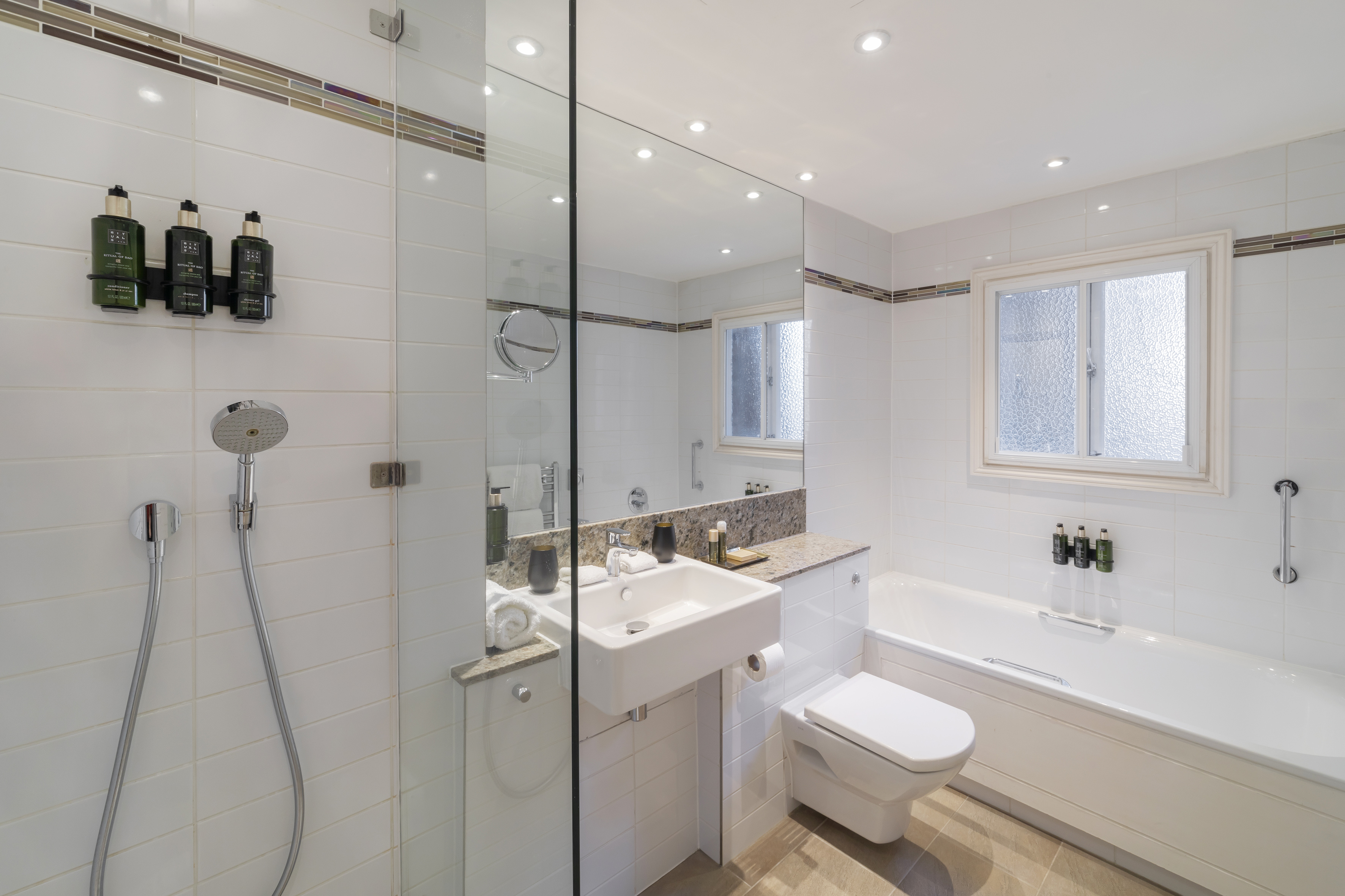 Modern white tiled hotel bathroom with shower, bathtub, sink, and toilet.