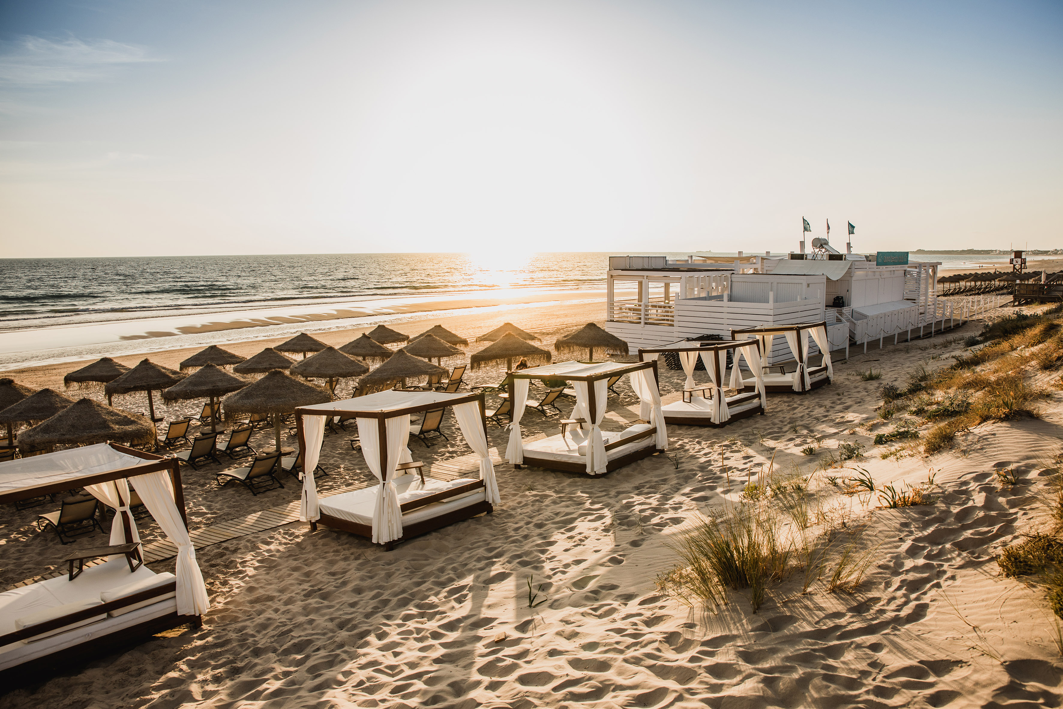 Luxury beach lounge at sunset with cabanas, straw umbrellas, and ocean.