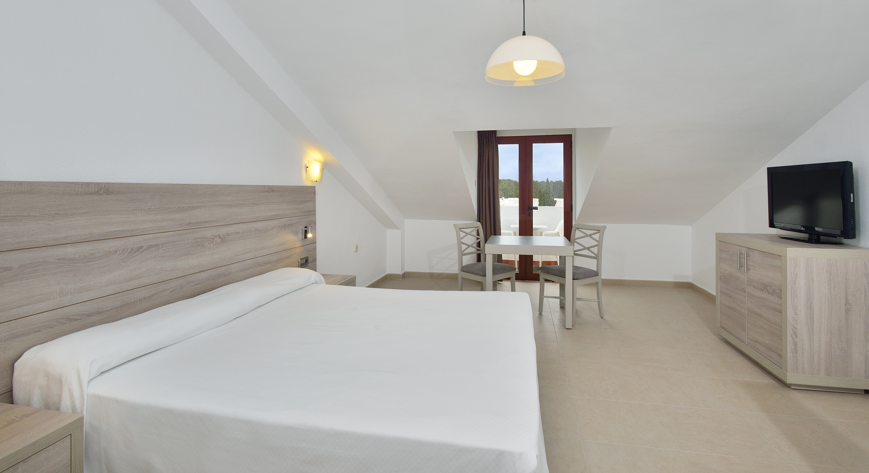 Hotel bedroom with white bed, wooden furniture, dining table, and balcony.