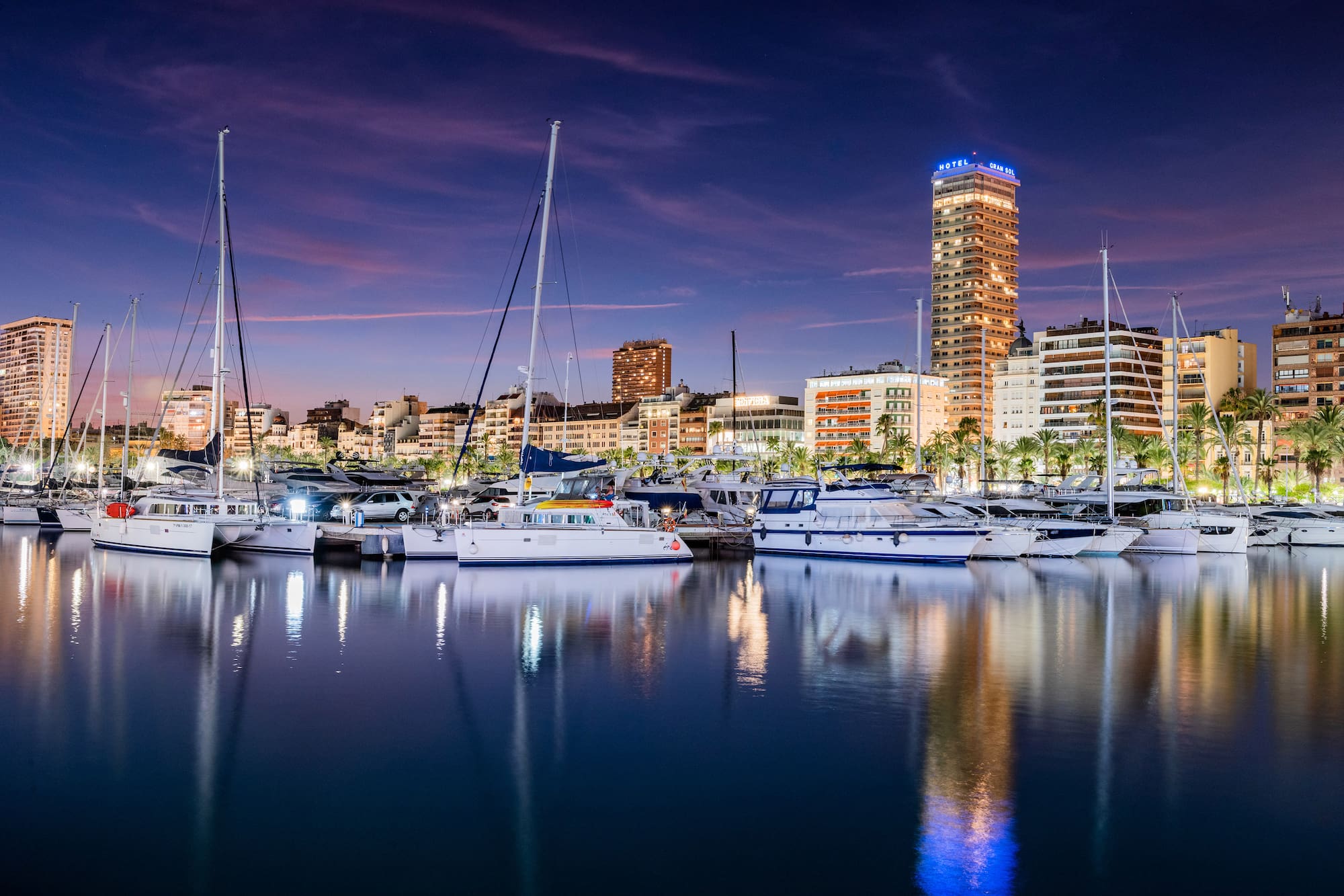 Alicante marina at dusk with yachts, illuminated city skyline, and reflections.