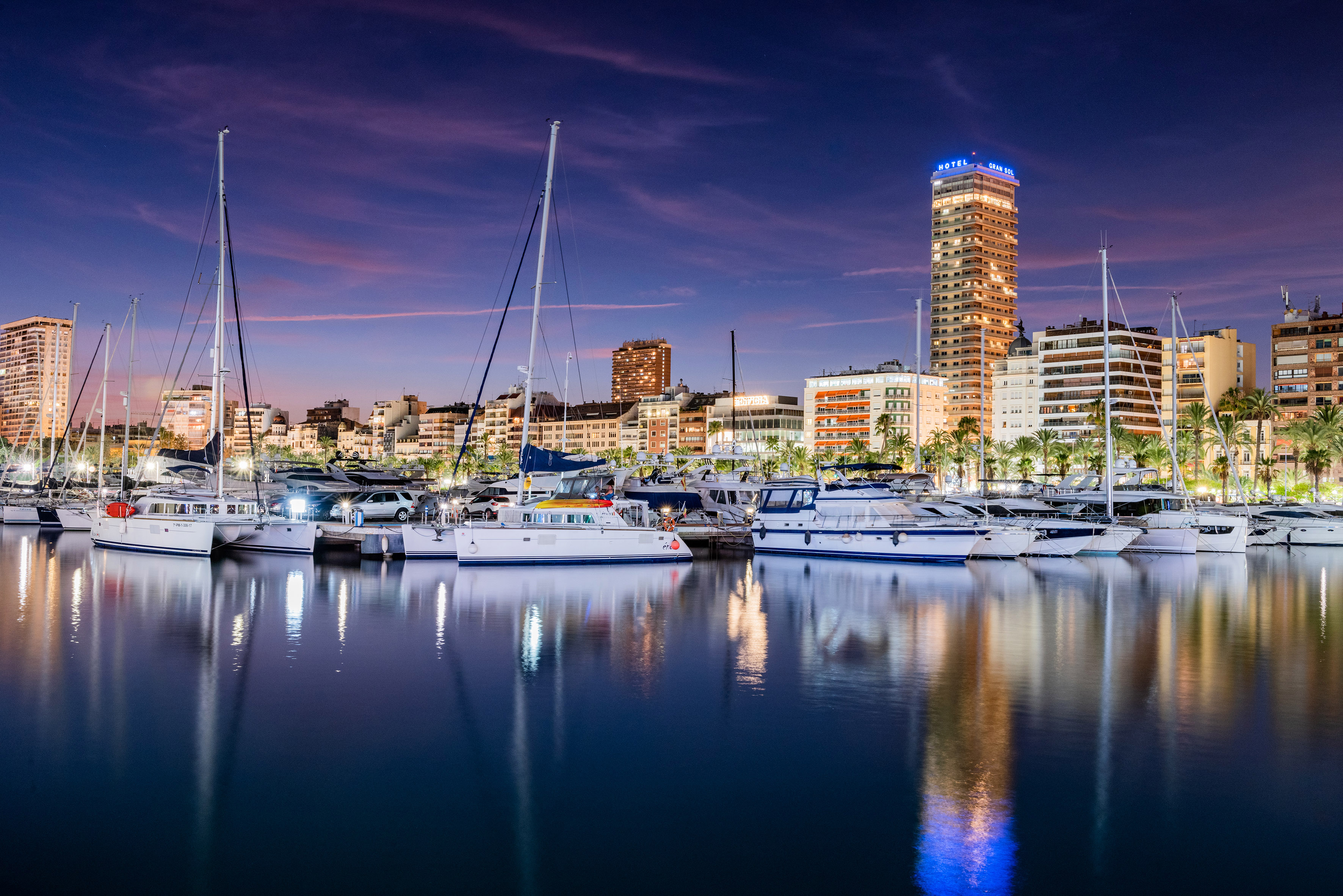 Alicante marina at dusk with yachts, illuminated city skyline, and reflections.