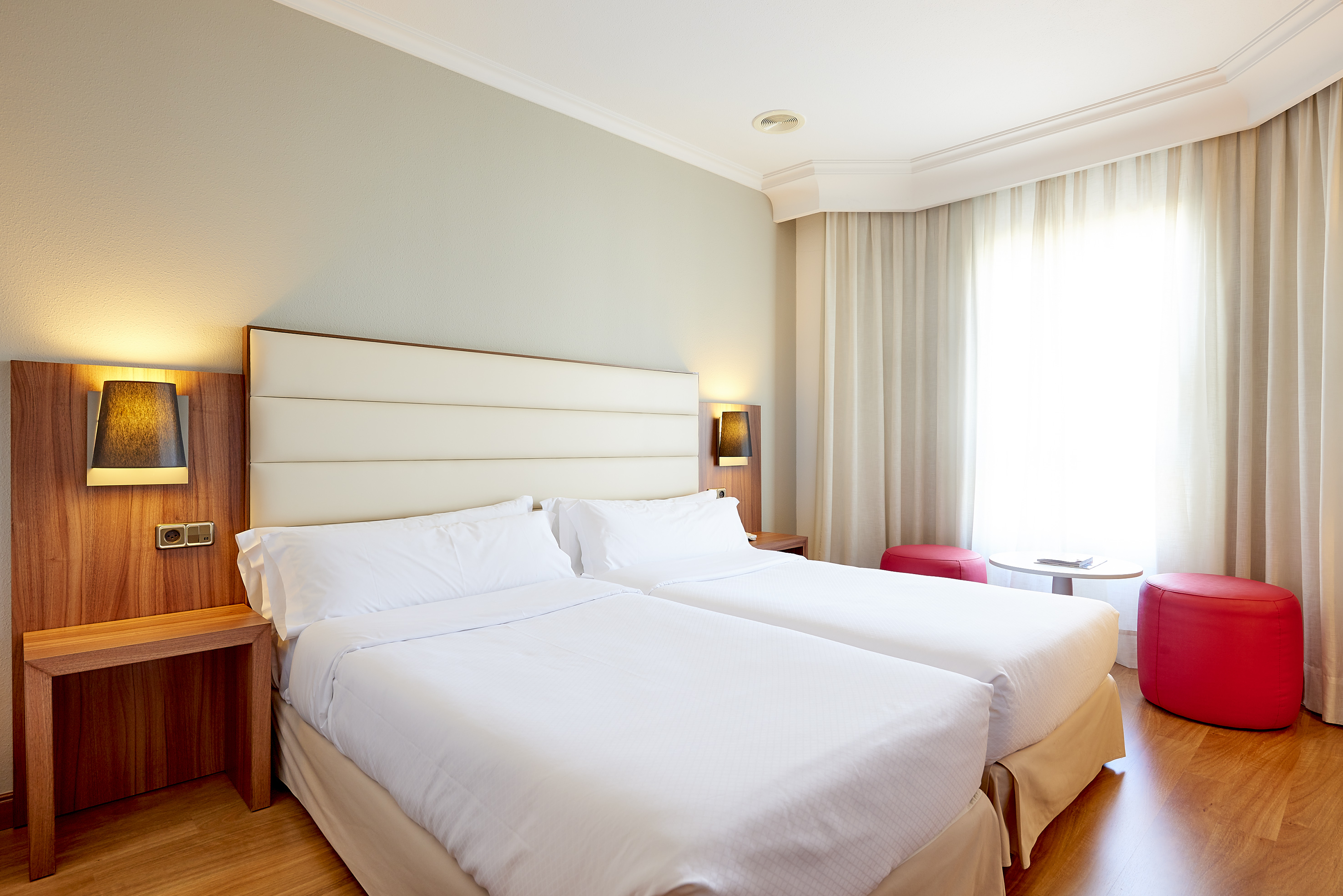Bright hotel room with twin beds, wooden floor, and red ottomans.