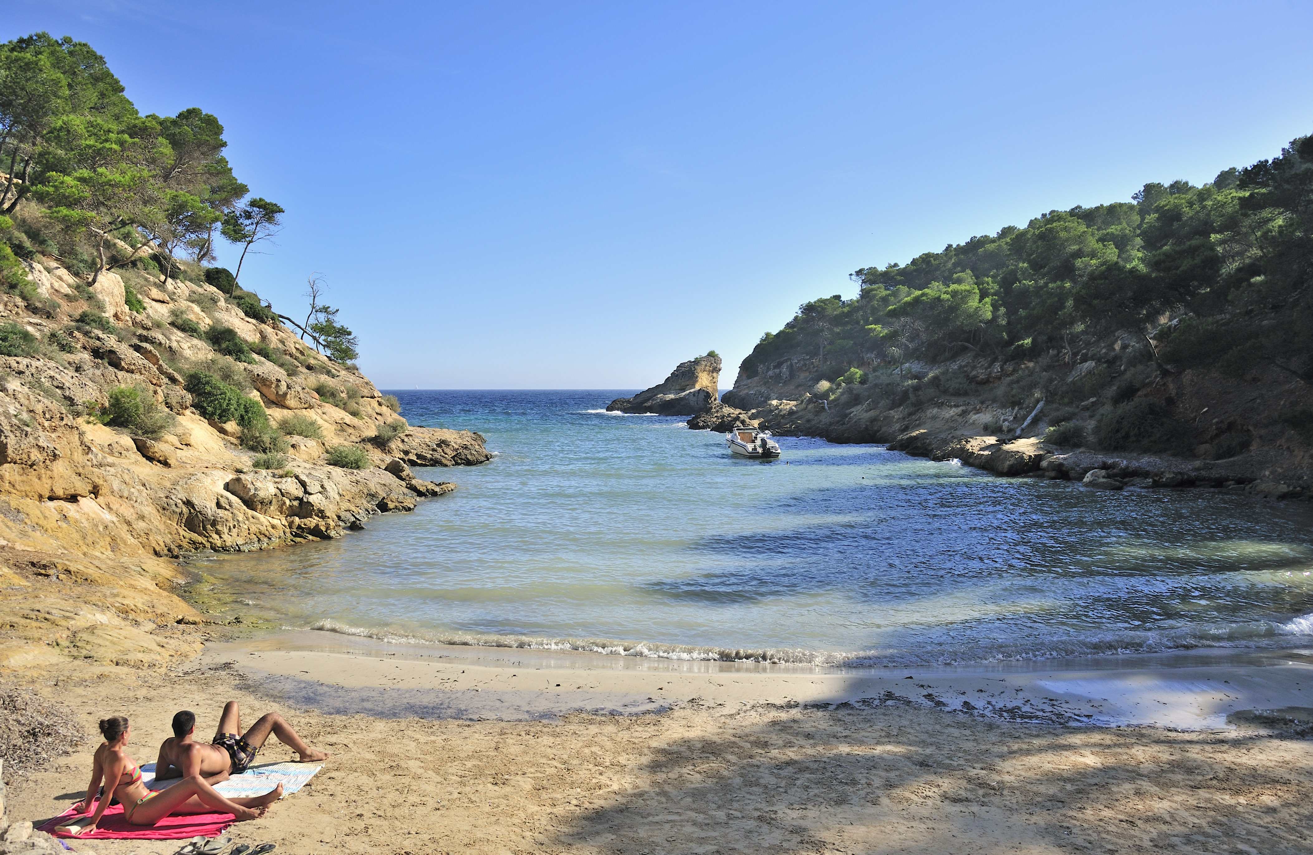 Cala mediterránea de arena, pareja, acantilados con pinos y bote en agua.