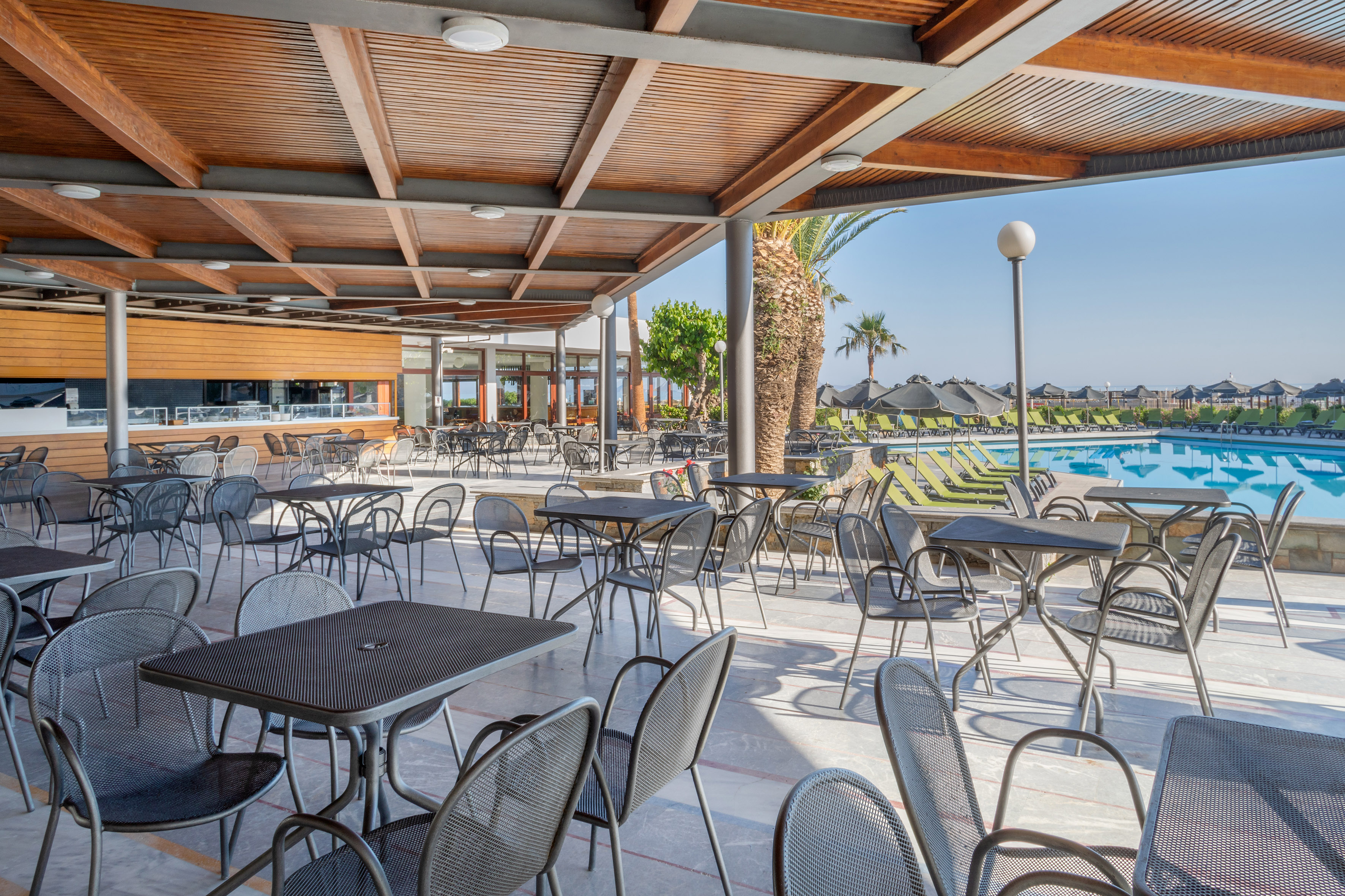 Sunny poolside restaurant terrace with outdoor seating and palm trees.