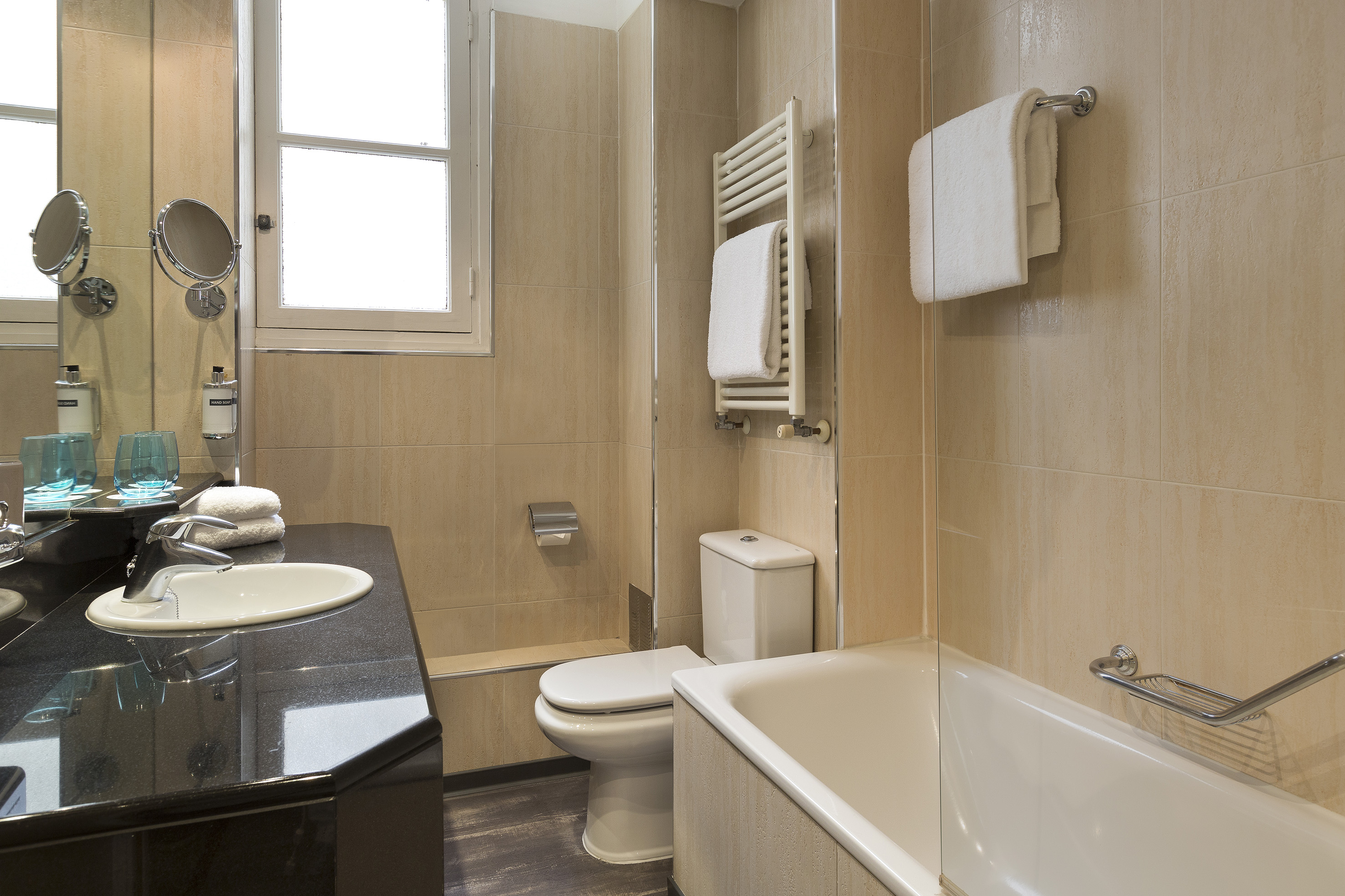 Beige tiled bathroom featuring black vanity, white tub, toilet, and window.