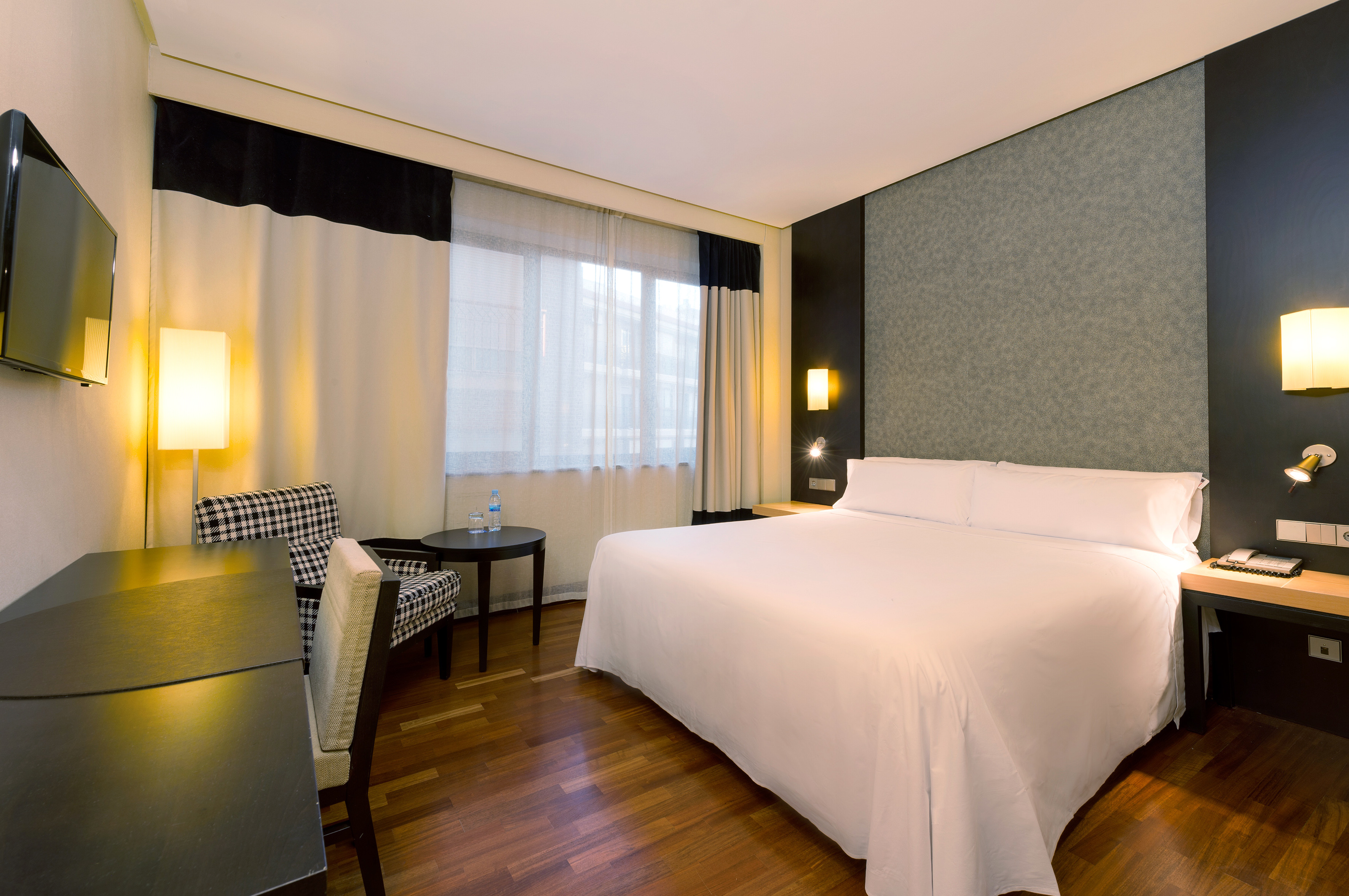 Modern hotel room with large bed, desk, armchair, TV, and window.